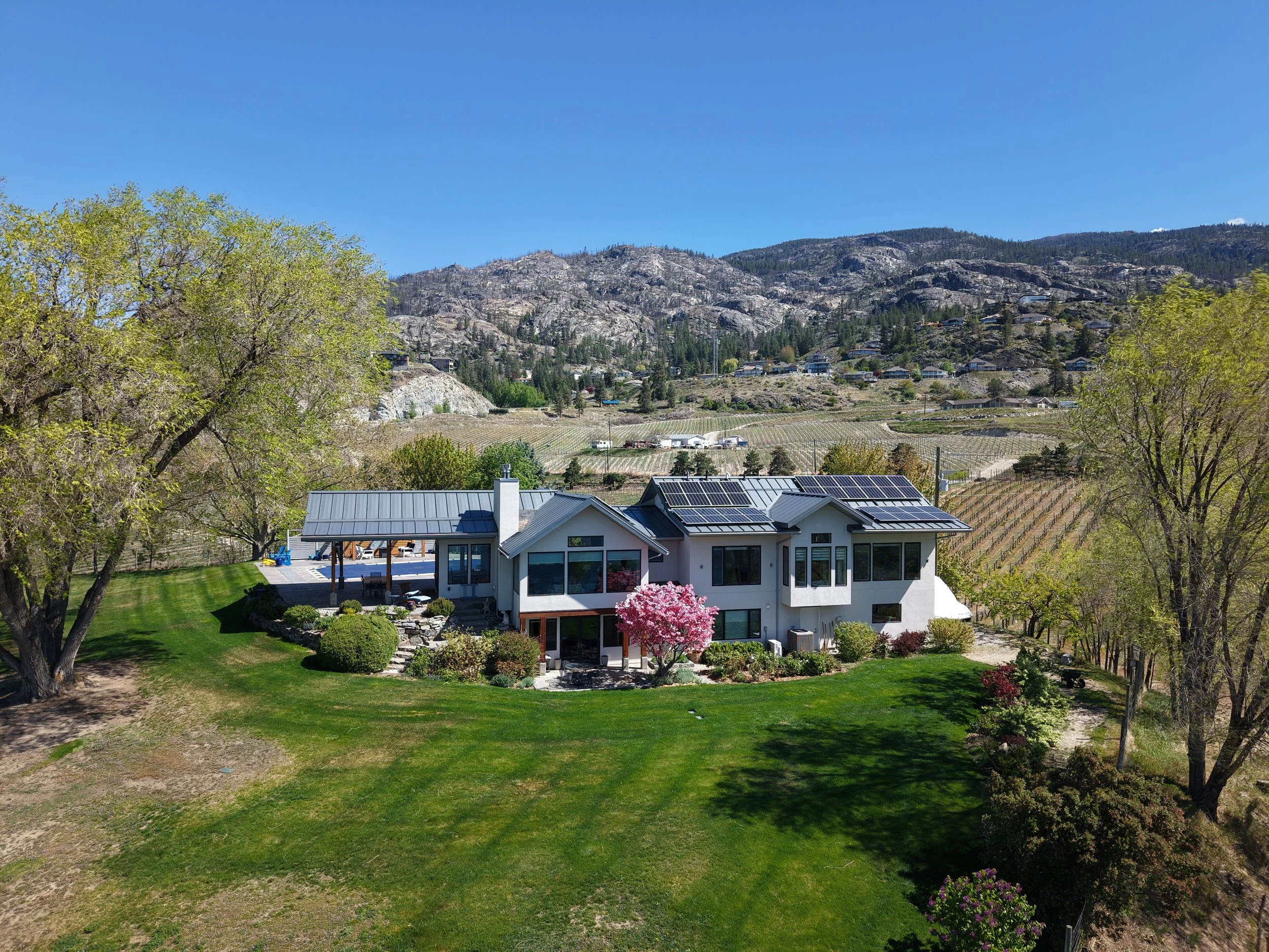 A modern house with solar panels on the roof, surrounded by a lush green lawn and flowering trees. In the background, there are hills with scattered houses and vineyards, and a clear blue sky overhead.