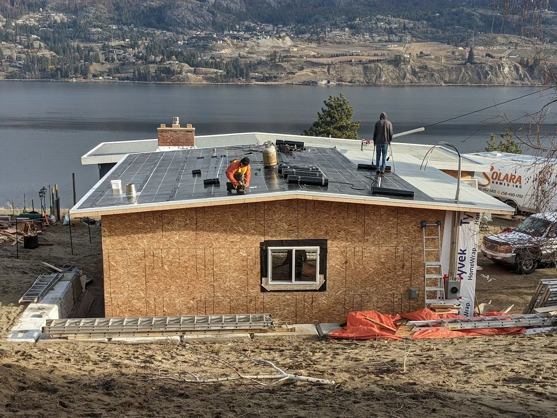 Construction workers installing a flat roof on a house by a lake, with a ladder and building materials around, large window on the wall.
