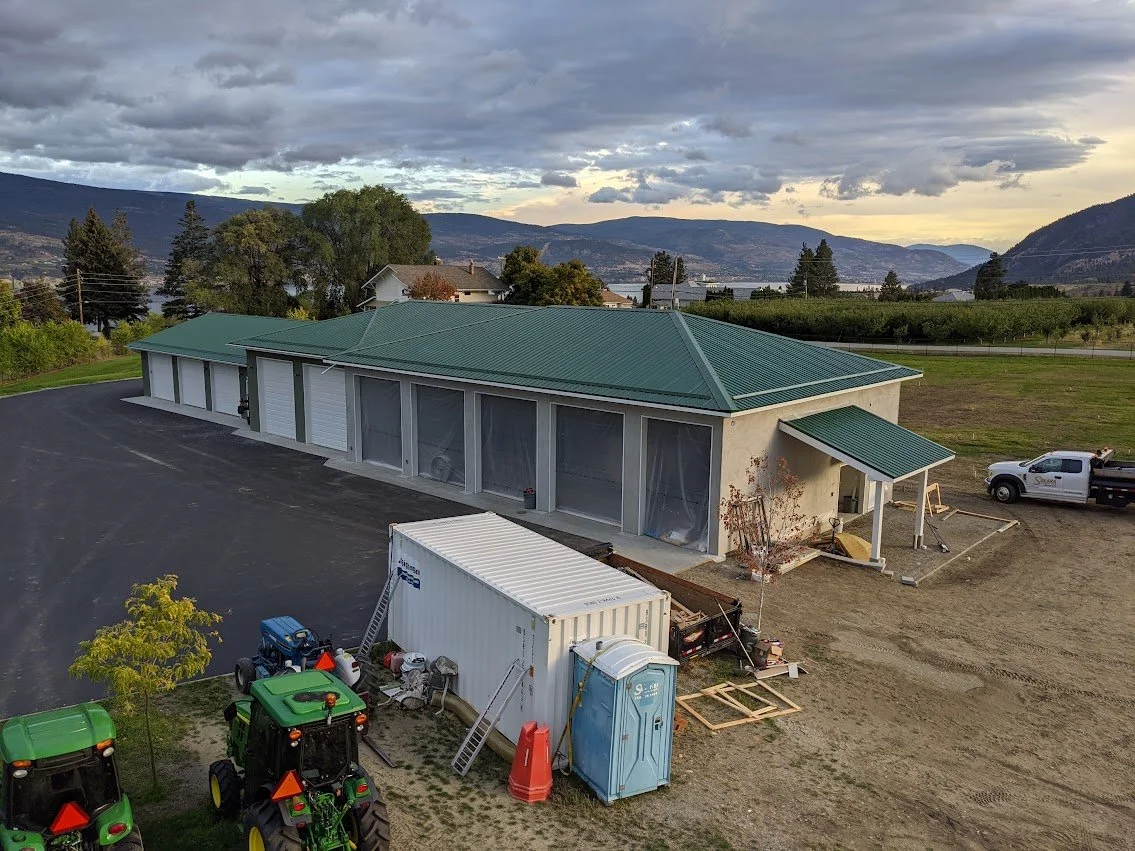 A large building with a green metal roof, multiple garage doors, and side windows, situated in a rural area with mountains in the background. There are construction supplies, two tractors, a portable toilet, and a shipping container in the foreground