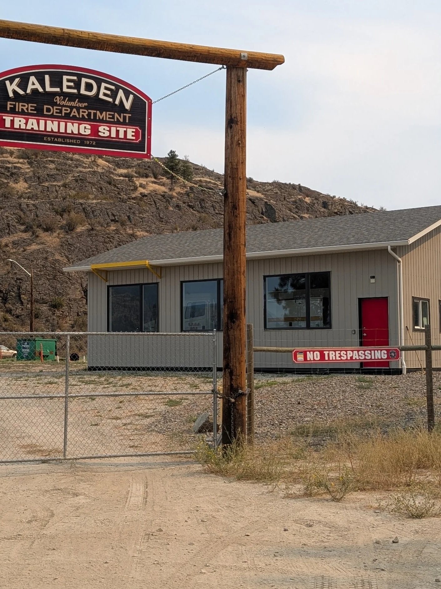 A fire training site sign, a building with large windows, chain-link fence with a 'No Trespassing' sign, desert terrain, rocky hillside background, and a wooden utility pole in front.