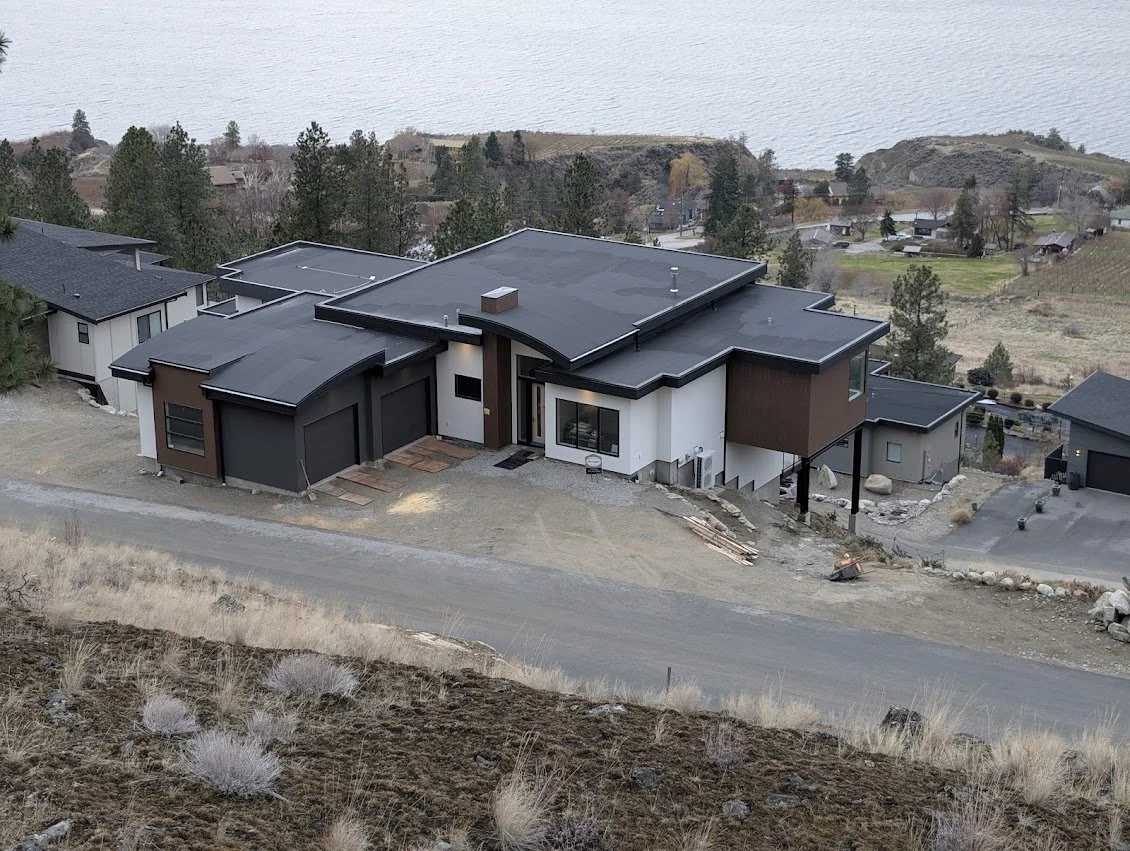 A modern house under construction on a hillside, with a black roof, white and brown exterior walls, and a gravel driveway, overlooking a rural landscape with trees and fields.