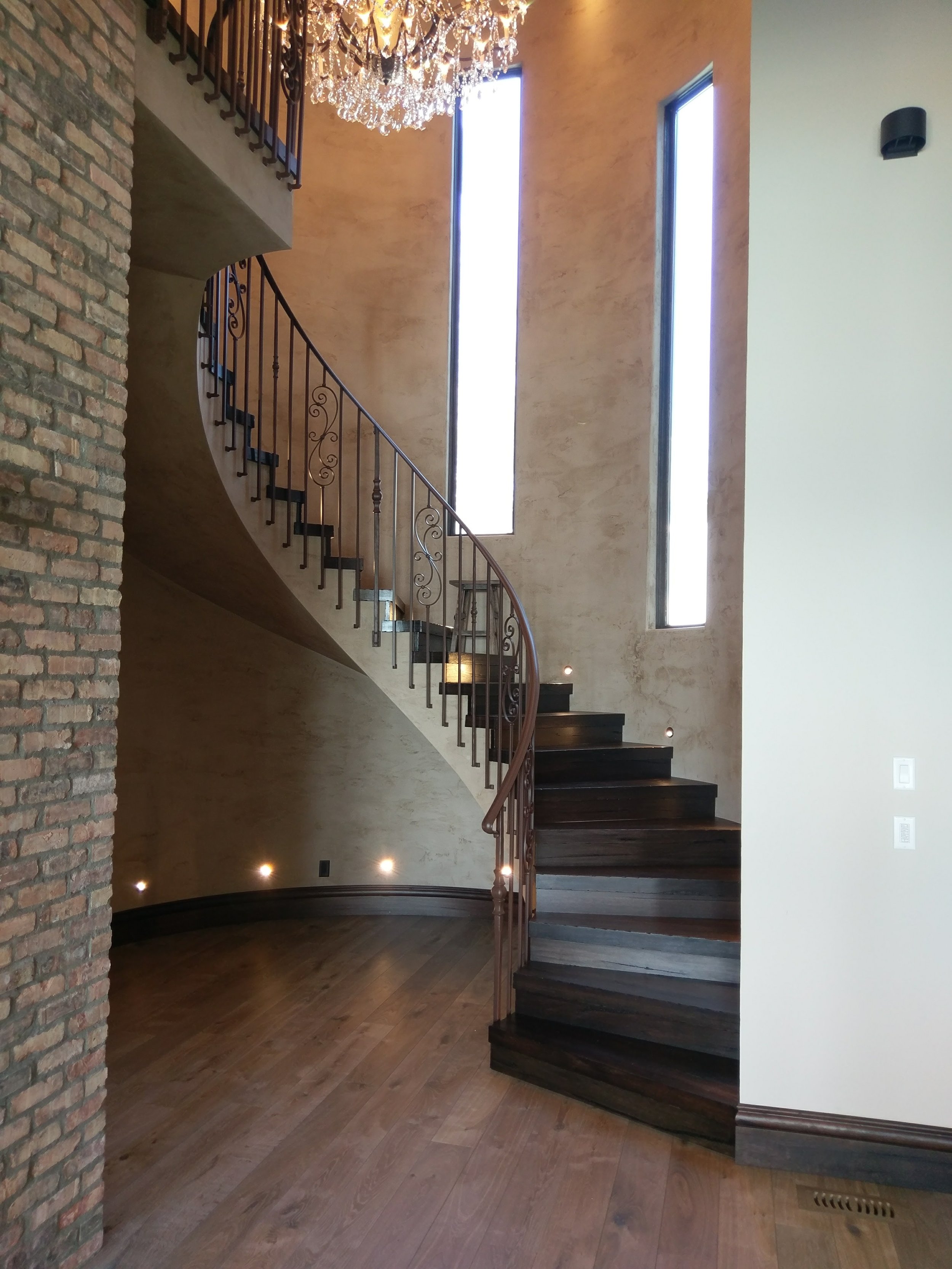 Interior view of a modern staircase with dark wood steps, iron railing, natural light from tall narrow windows, a brick wall on the left, and a large ornate chandelier hanging from the ceiling.