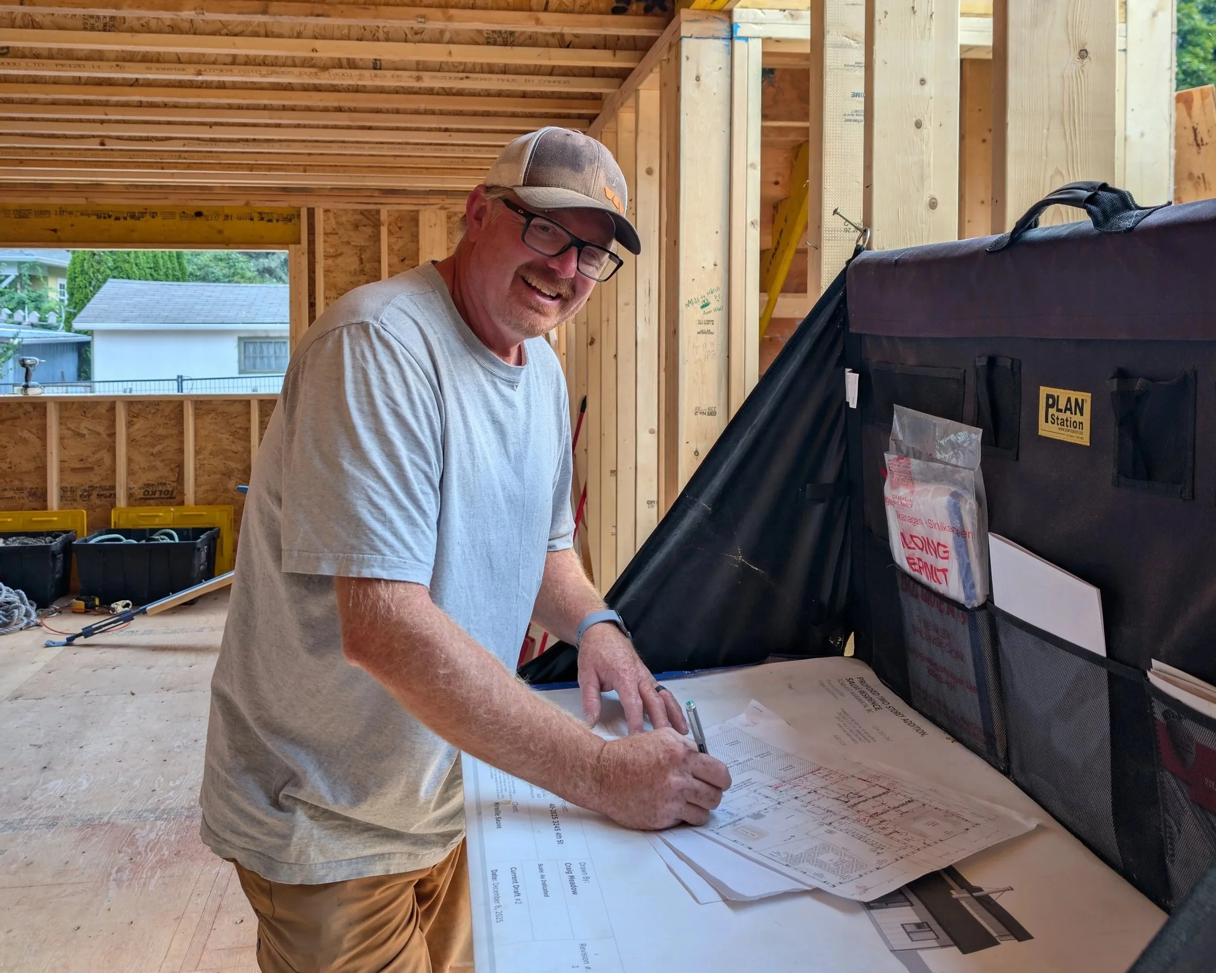A man wearing glasses and a cap is working on blueprints at a construction site with wooden framing.