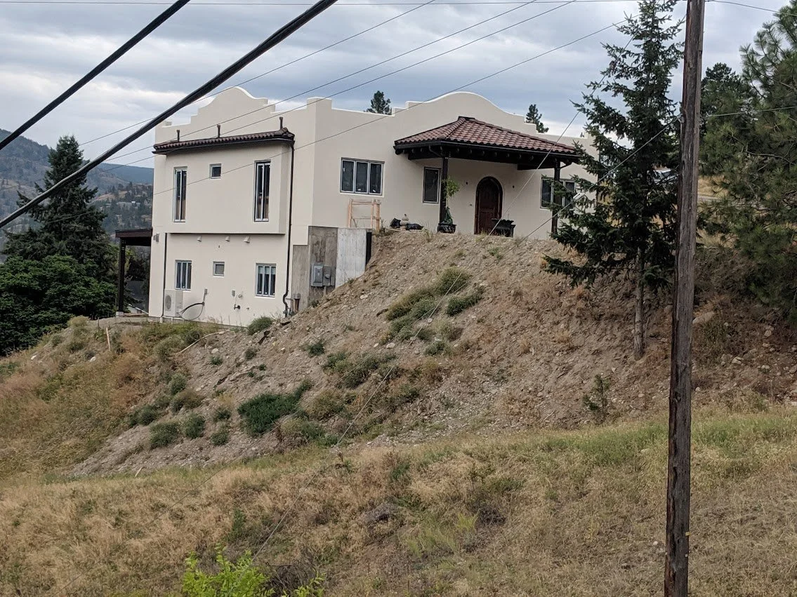 A two-story house on a hillside with a dirt slope in front, surrounded by trees and power lines, under a cloudy sky.