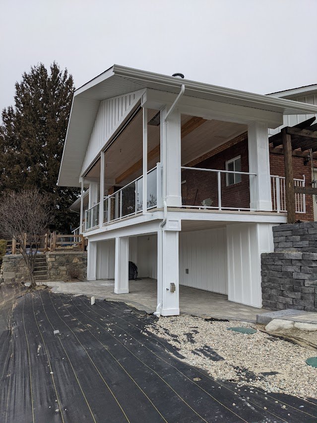 Front view of a raised house with white siding, a balcony with glass railing, and an under-building space with ground covering.