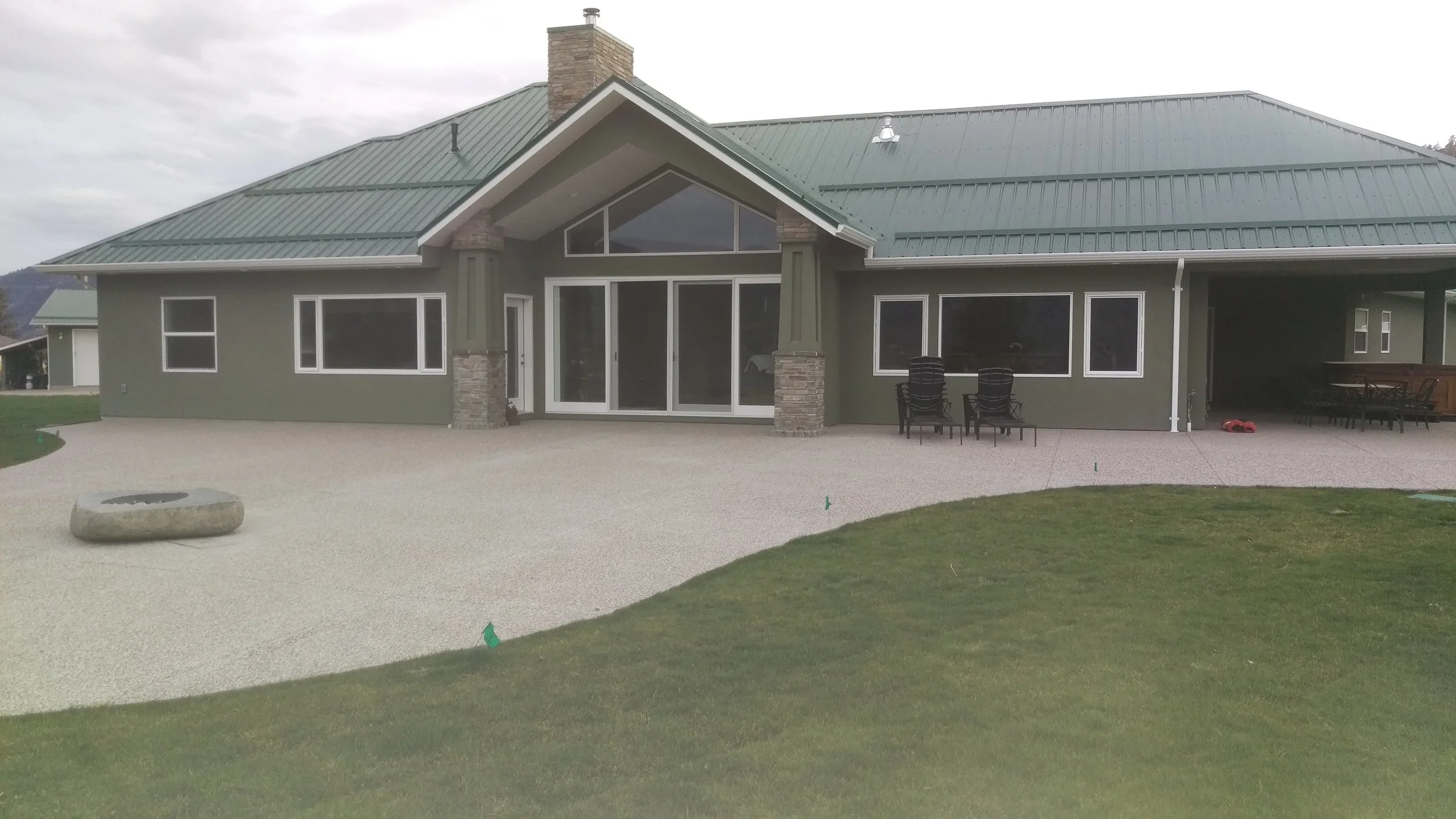 Behind a house with a green metal roof, there is a spacious gravel patio with seating and a small grassy area.