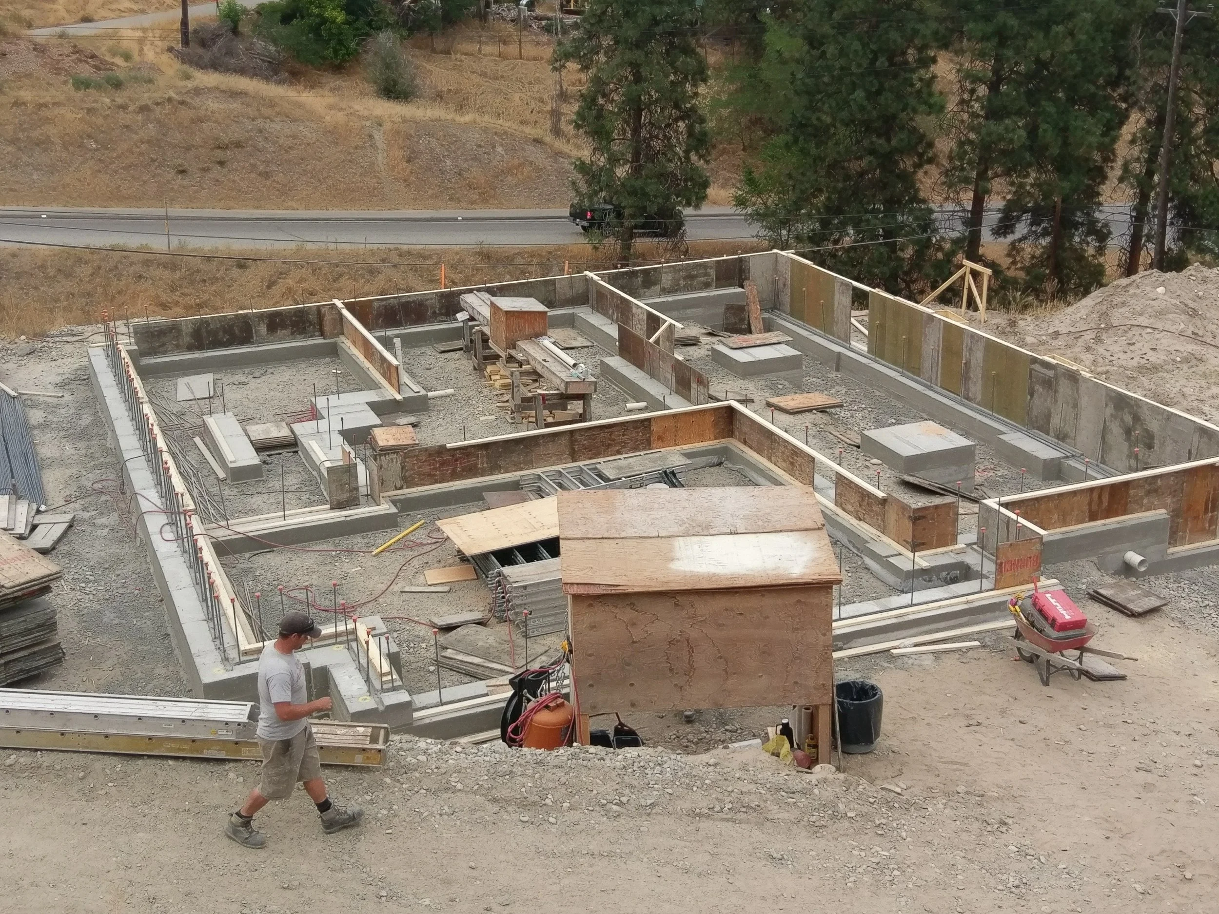 Construction site with concrete foundation and framework being built. Two workers are seen, one in a white t-shirt and beige shorts, and the other in a white shirt and gray pants. Trees and a roadway are visible in the background.
