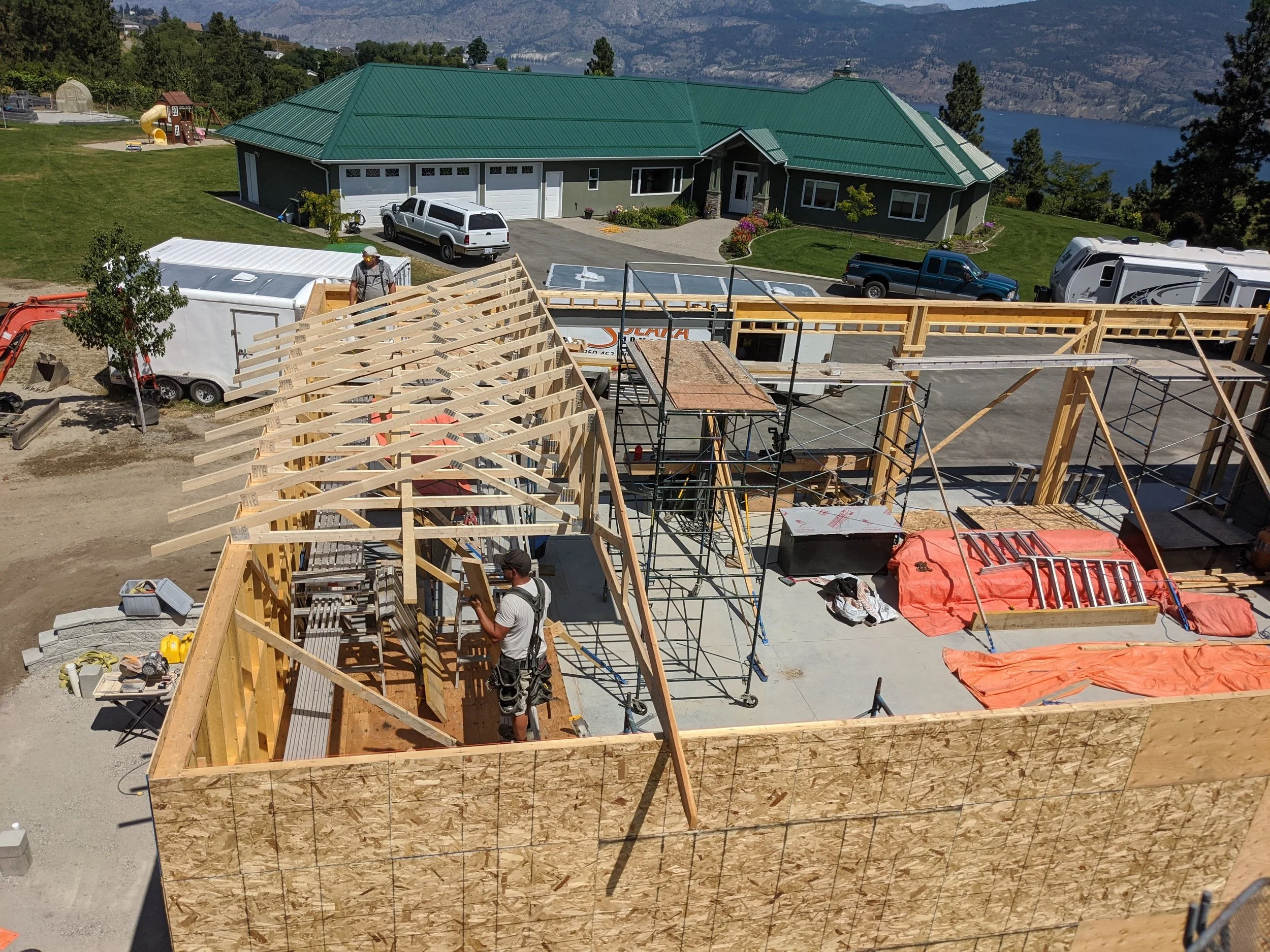 Construction site with framing for a building, workers, and construction equipment, overlooking a house and lake in the background.