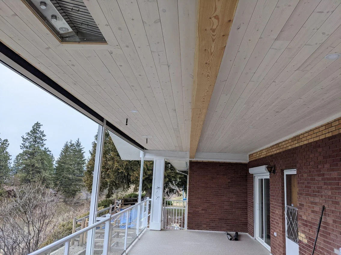 Front porch of a house with a new unfinished ceiling, brick wall, sliding glass door, and railing. Trees and cloudy sky visible outside.