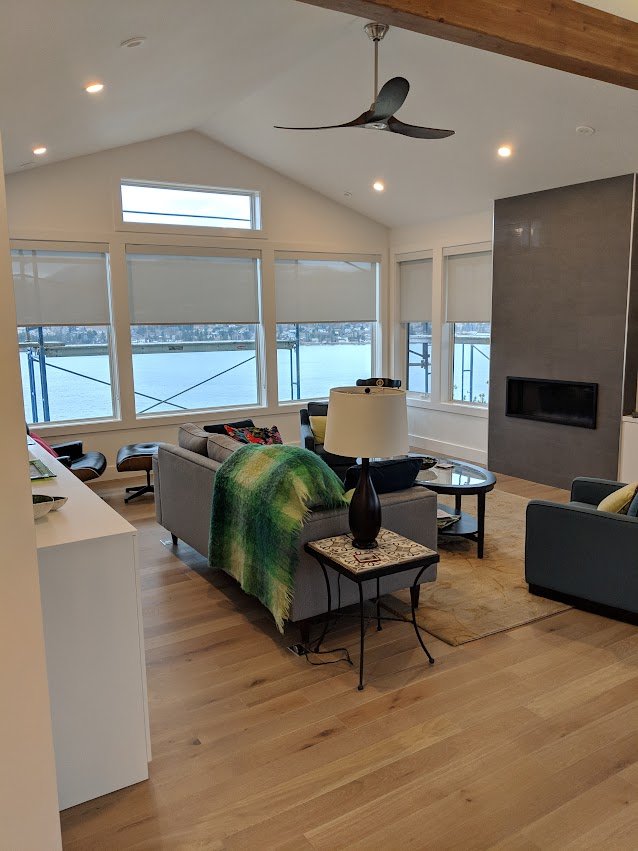 Living room with large windows overlooking water, modern fireplace, ceiling fan, and cozy seating area with throw blanket.