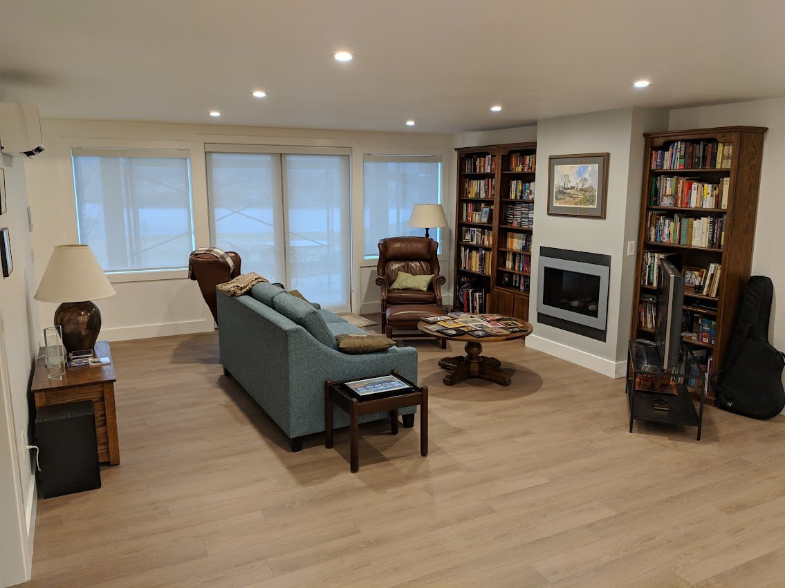 A cozy living room with wooden flooring, a blue sofa, leather armchairs, wooden bookshelves filled with books, a window with closed blinds, a fireplace, and lamps for lighting.