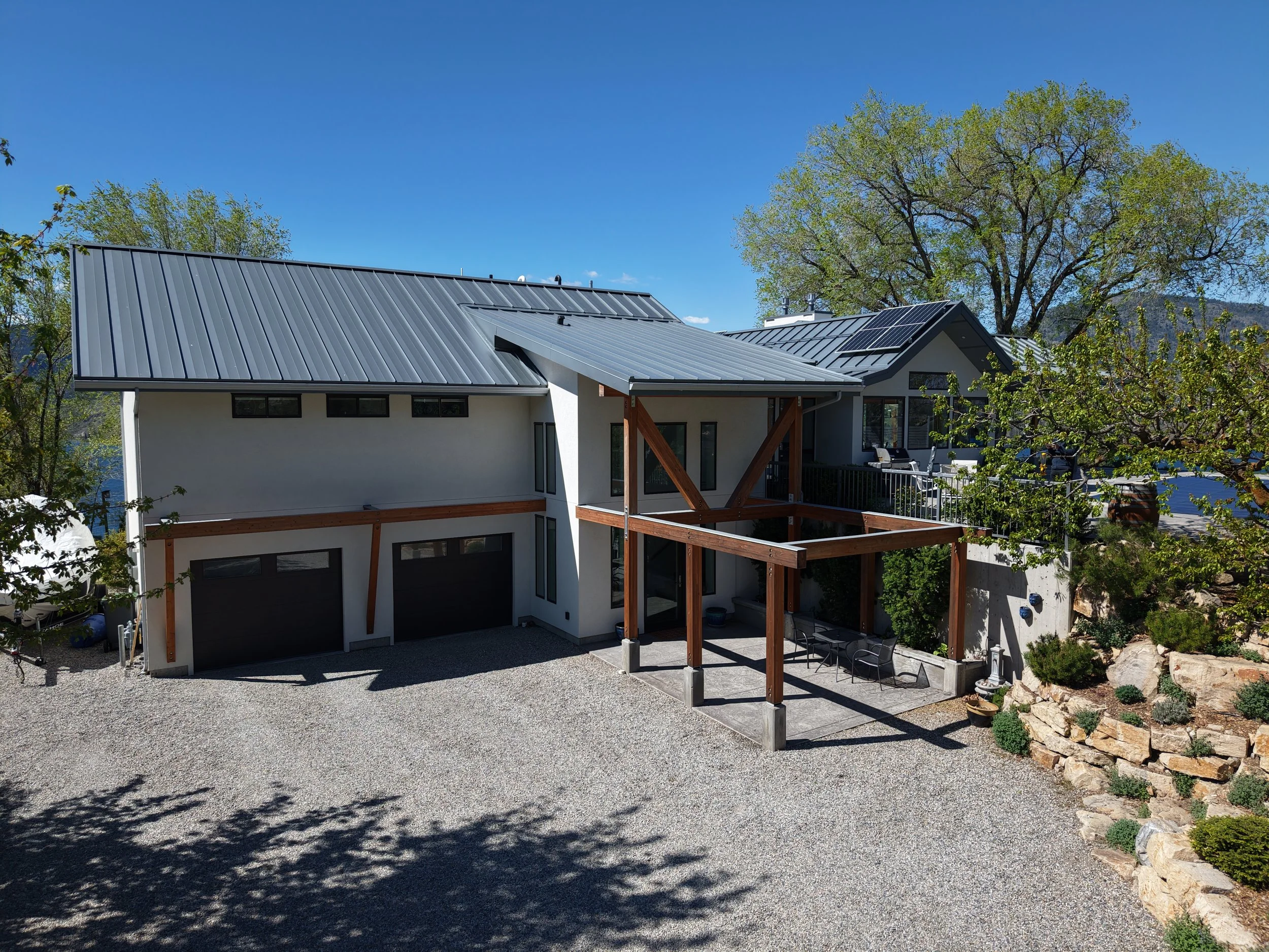Modern house with metal roof, outdoor patio area with seating, landscaped yard, and large trees under a clear blue sky.
