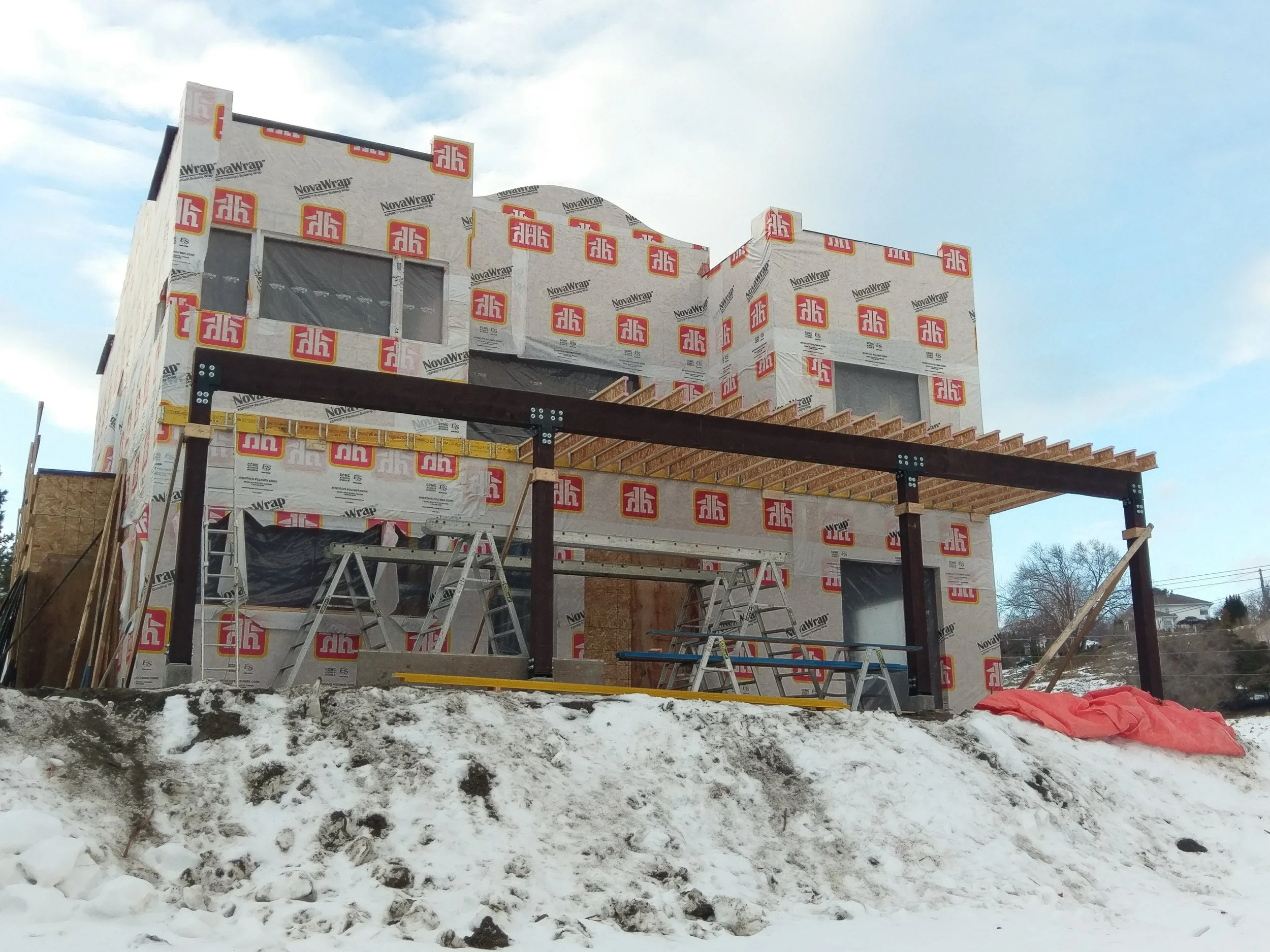 A house under construction with scaffolding, wrapped in NovaWrap weather barrier, with snow on the ground and a partly cloudy sky.