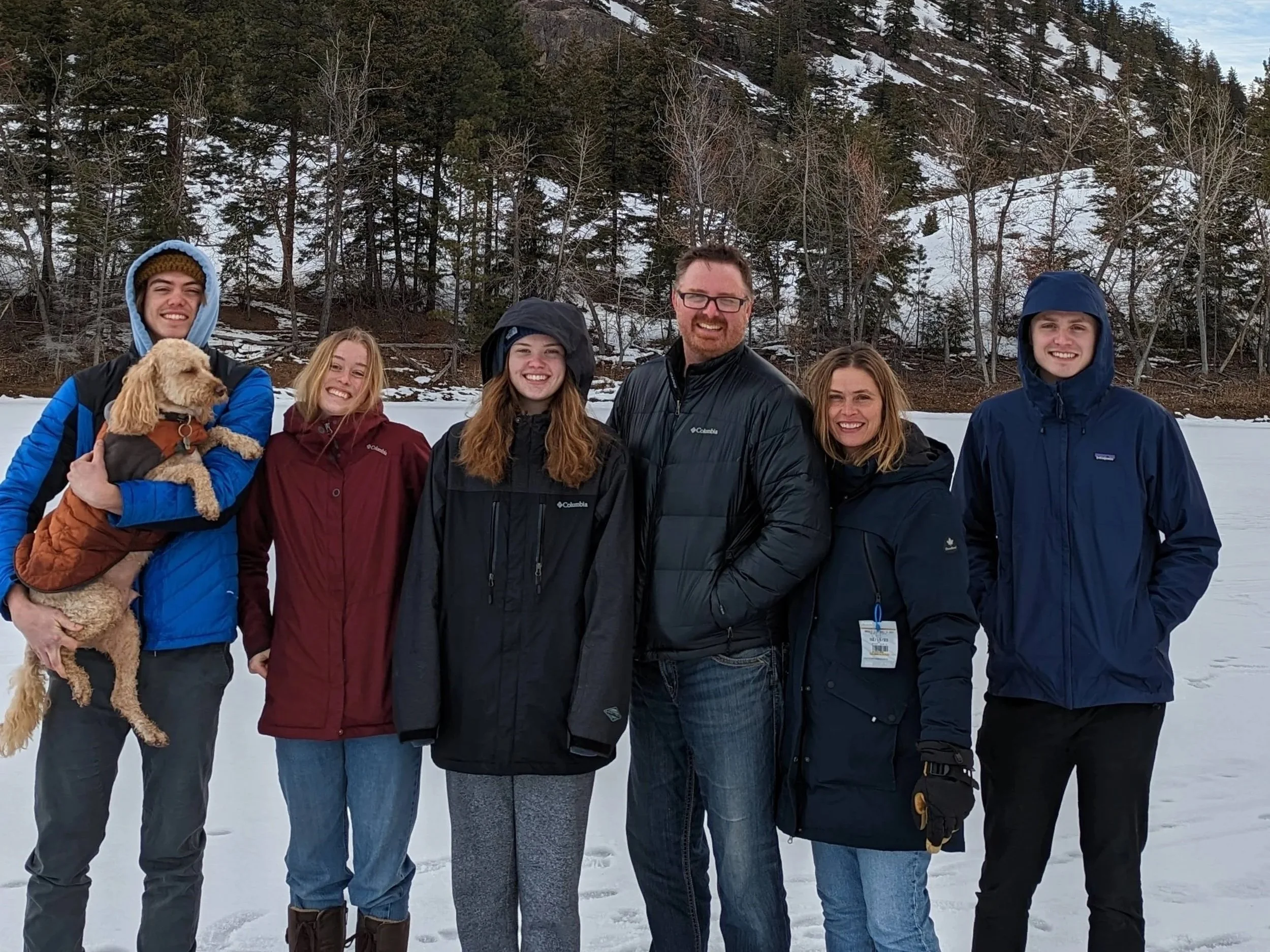 Group of six people standing on snow-covered ground in a winter landscape with trees and hills in the background, posing for a photo.