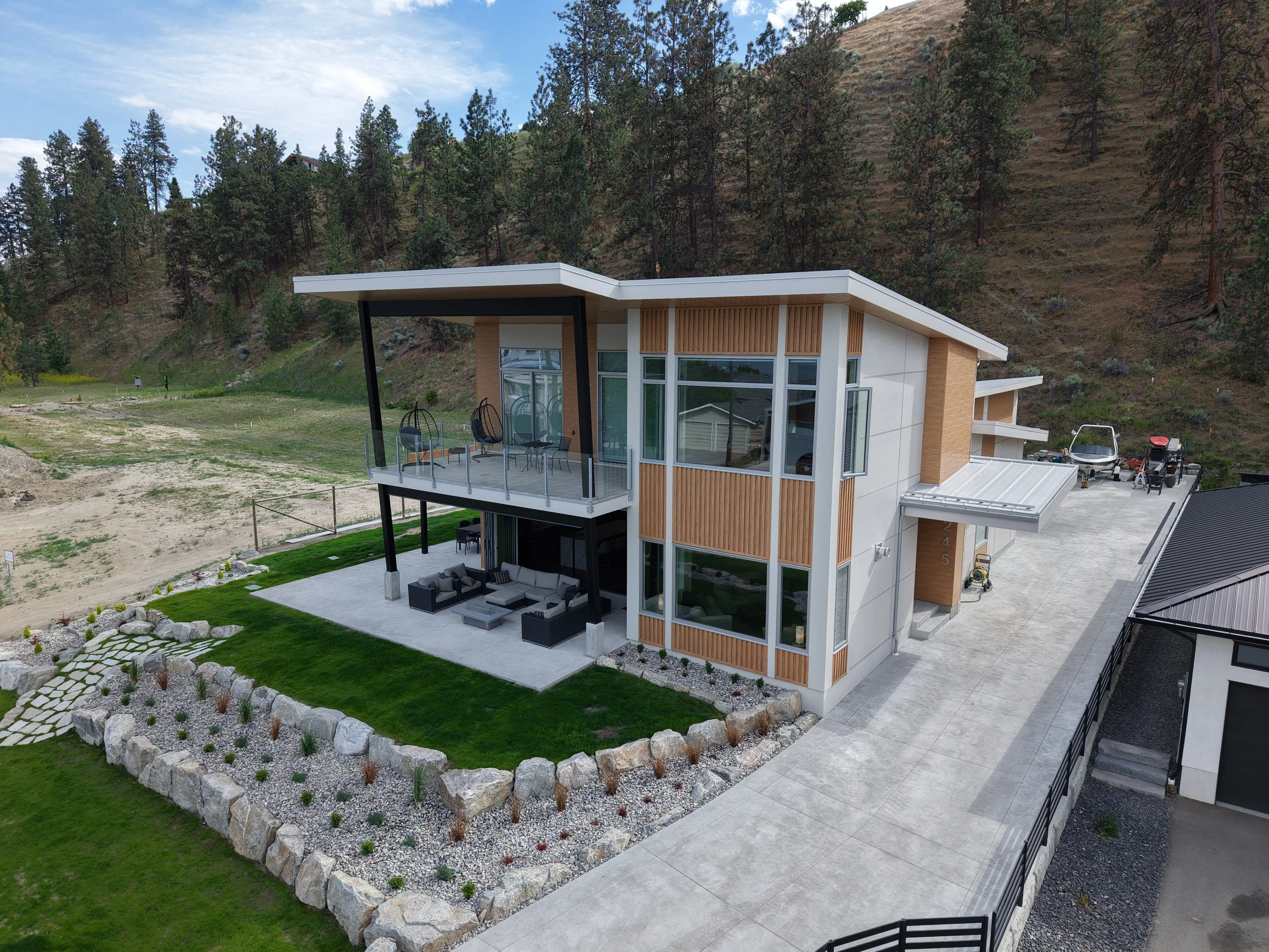 Modern house with large windows and a spacious balcony overlooking a landscaped yard with rocks and plants, set against a hillside with trees.
