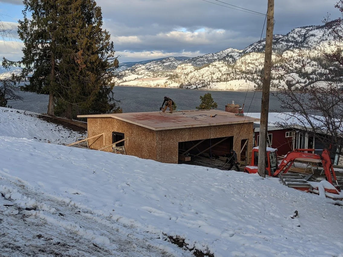 A worker on a flat rooftop under construction, with snow on the ground and a body of water and snow-covered mountains in the background.