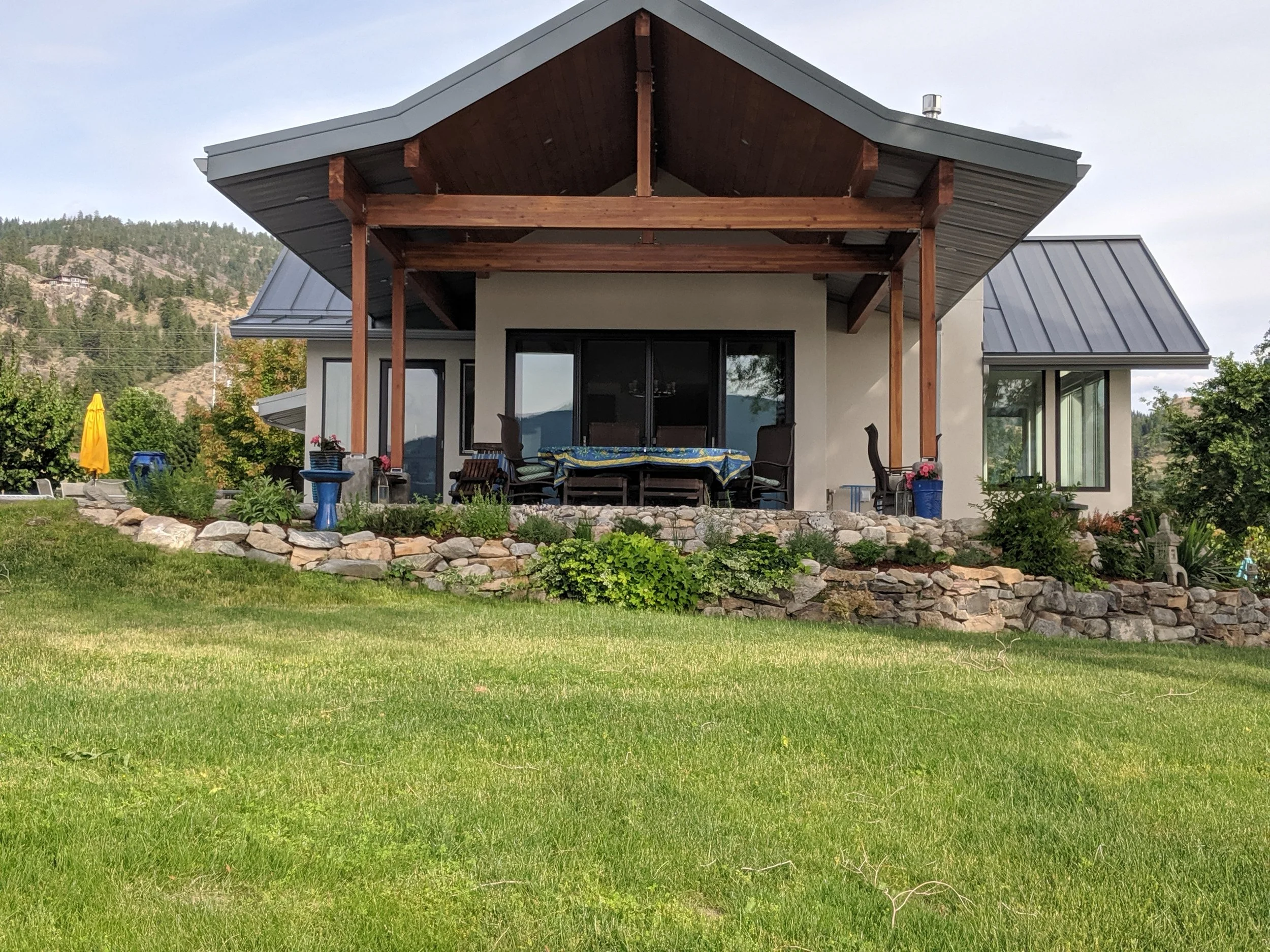 View of a modern house with a covered patio area, outdoor furniture, a garden, and a mountainous landscape in the background.