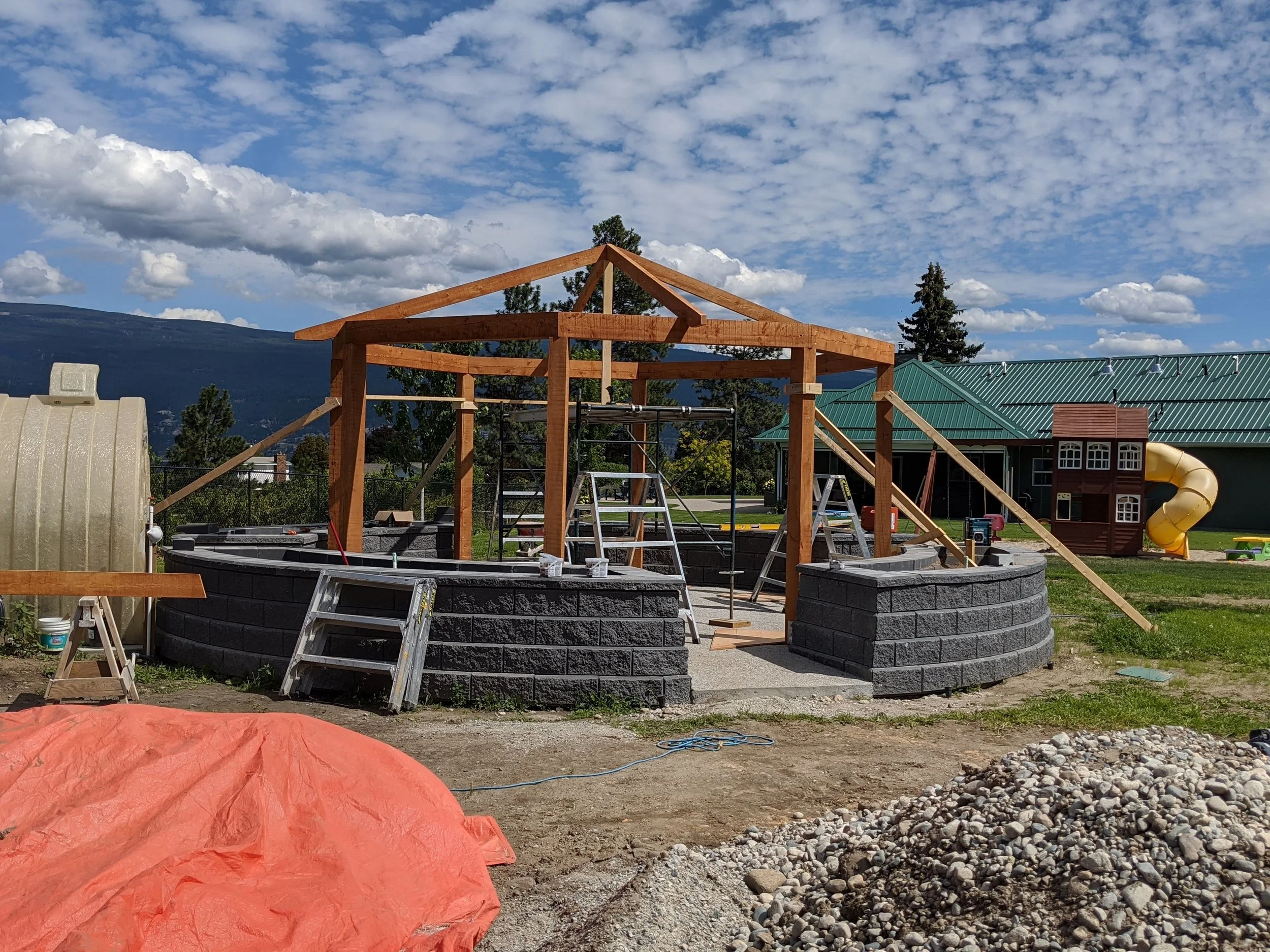 A house under construction with a wooden frame, scaffolding, and a partially built stone wall, set against a backdrop of blue sky with fluffy clouds and distant mountains.