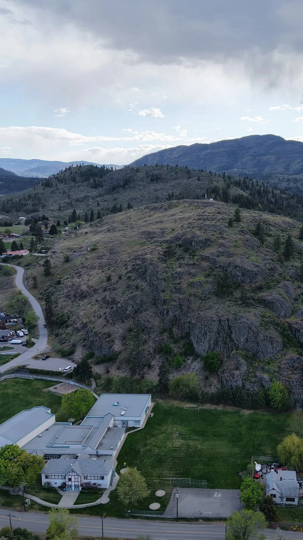 A hilly landscape with rocky terrain, sparse trees, and a small residential area at the base. The sky is cloudy.