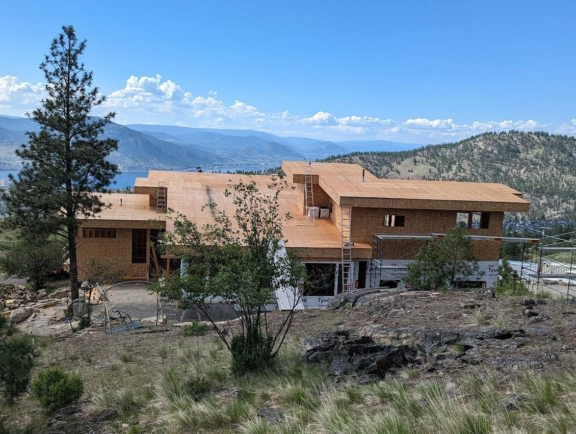 House under construction on a hillside, surrounded by trees with mountains and a lake in the background, blue sky with clouds.