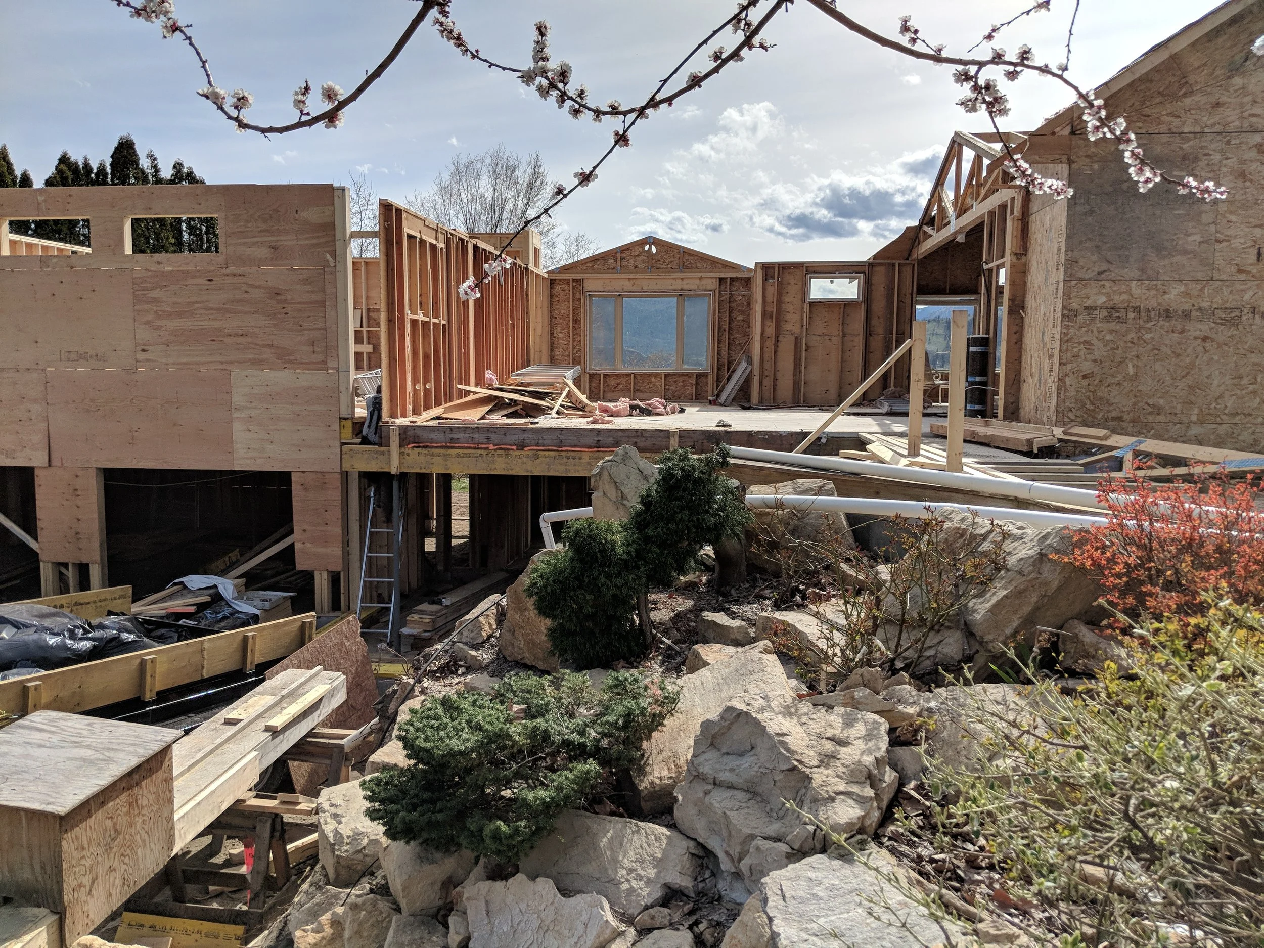 Under construction view of a house with wooden framing, construction debris, rocks, and plants in the foreground, with a cloudy sky in the background.