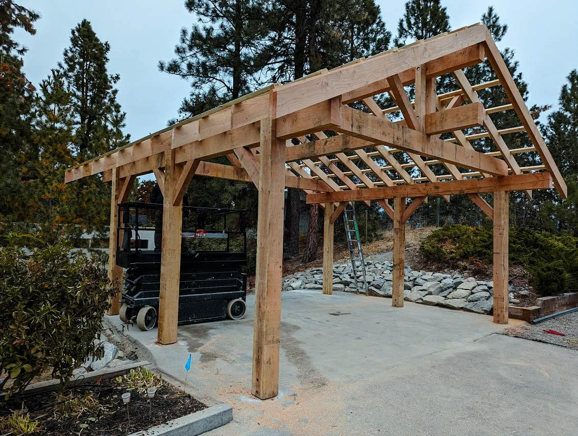 Wooden carport under construction in a backyard with trees and rocks, including a ladder and a lift machine.