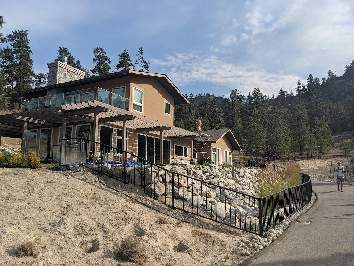 Modern house under construction on a hillside with scaffolding, rocks, pine trees, and two people near the house.