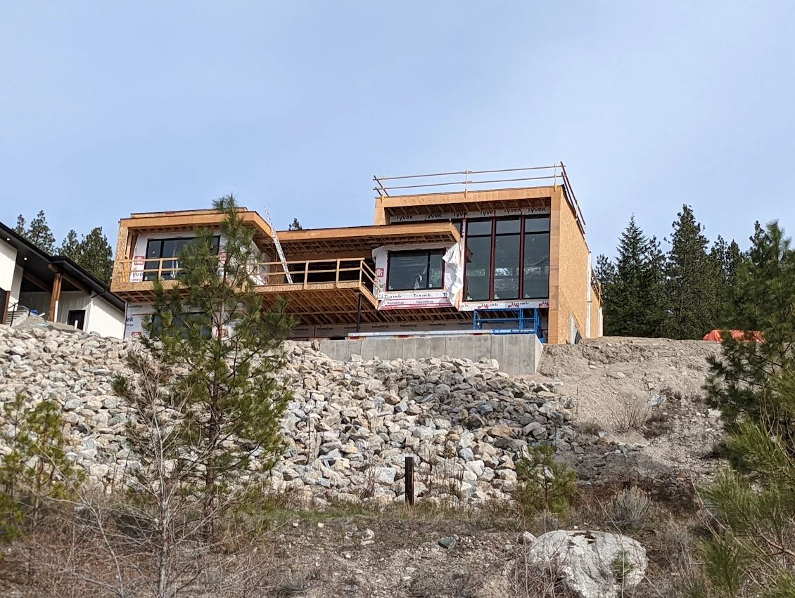 Modern house under construction on a hillside with trees in the background and construction materials around.