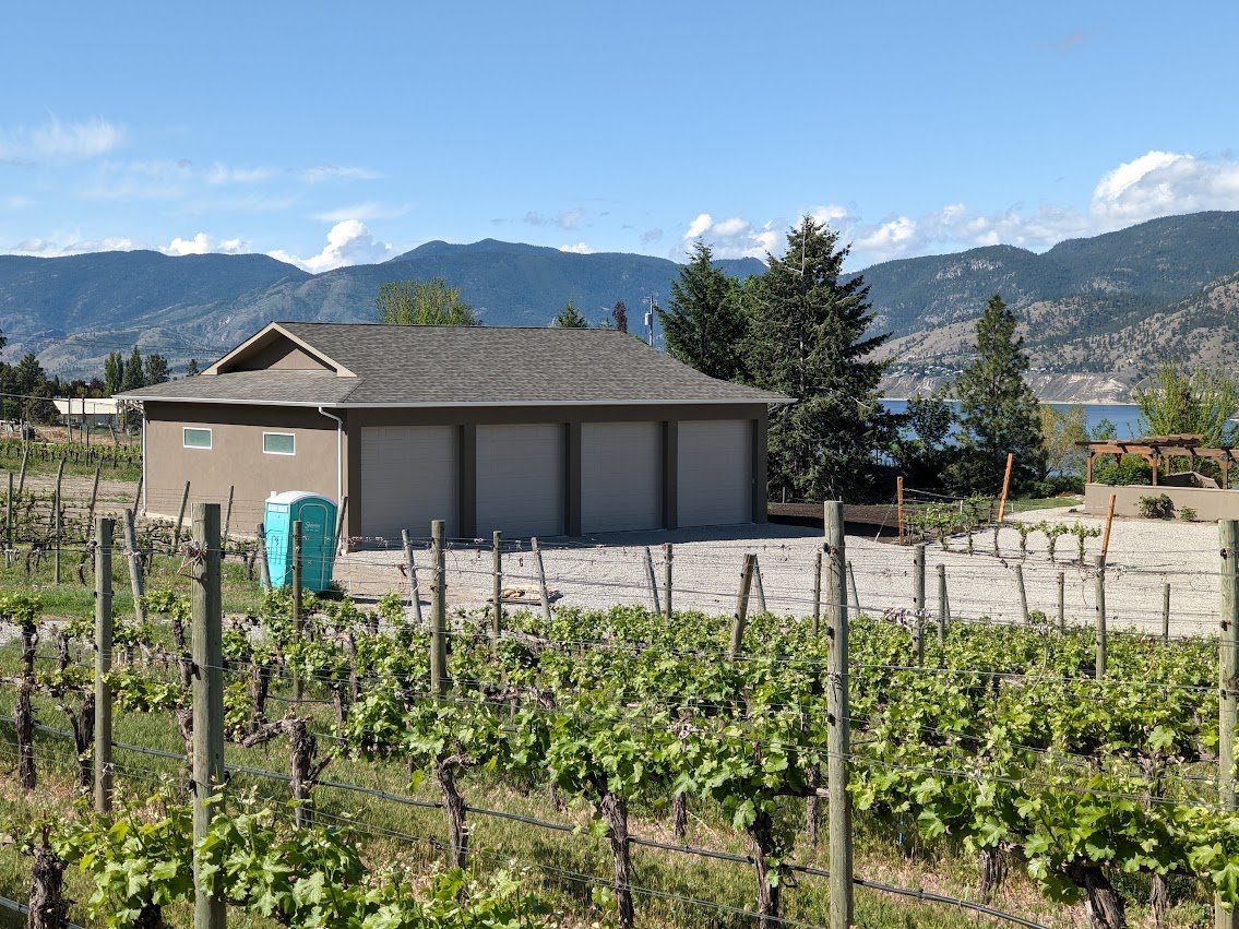 A vineyard with rows of grapevines, a small building with three garage doors, and a scenic mountain landscape in the background.