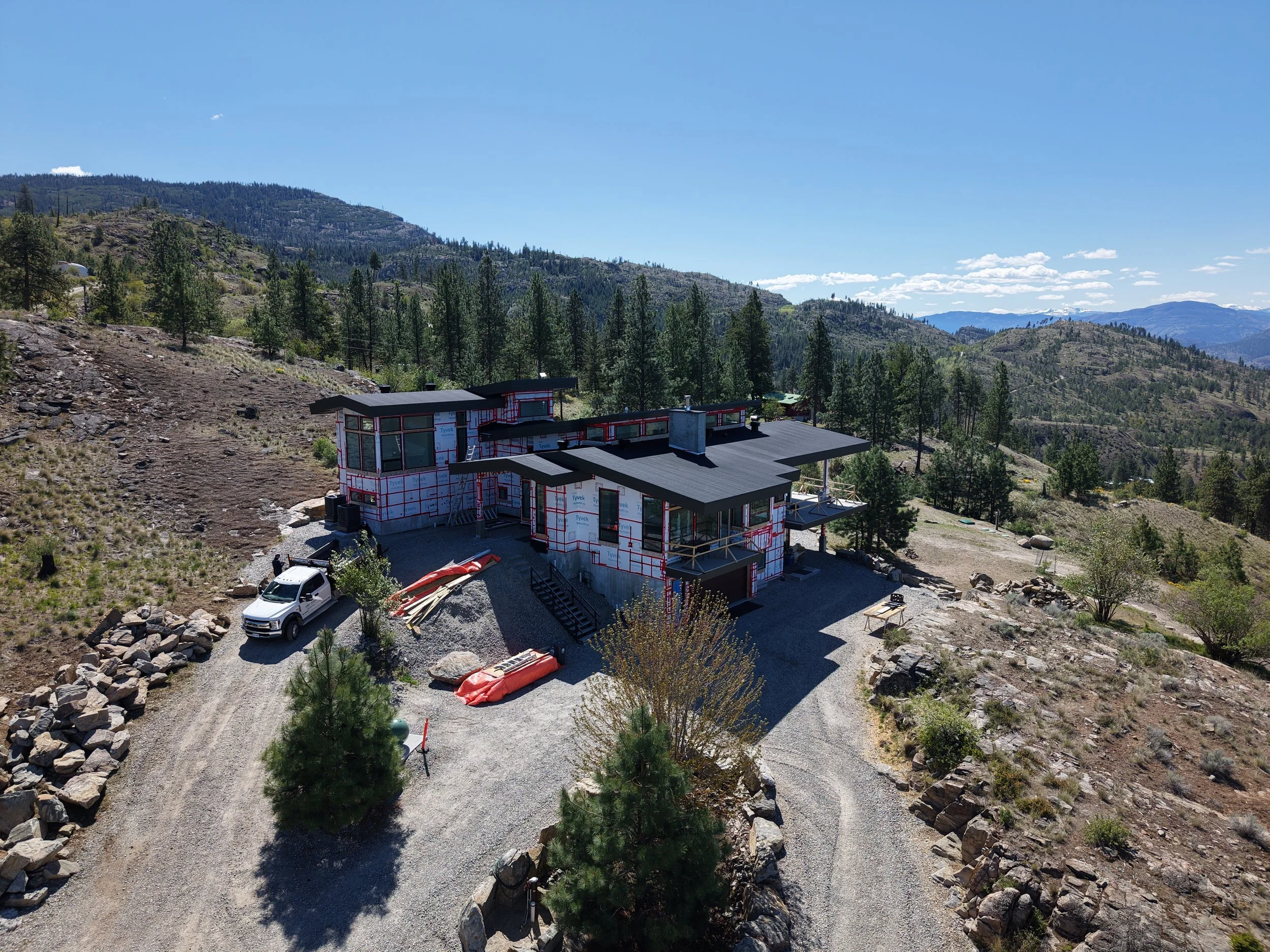A house under construction in a mountainous area with numerous trees and a clear blue sky.