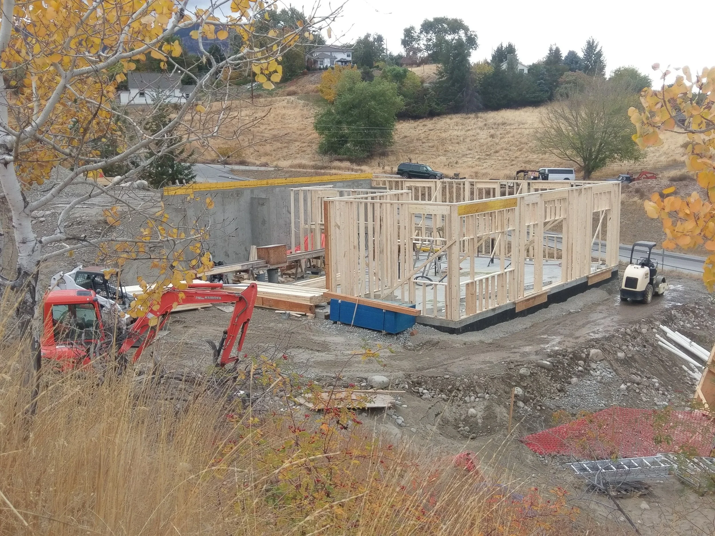 Construction site with wooden framing for a building, construction equipment, and vehicles, with trees and houses in the background.