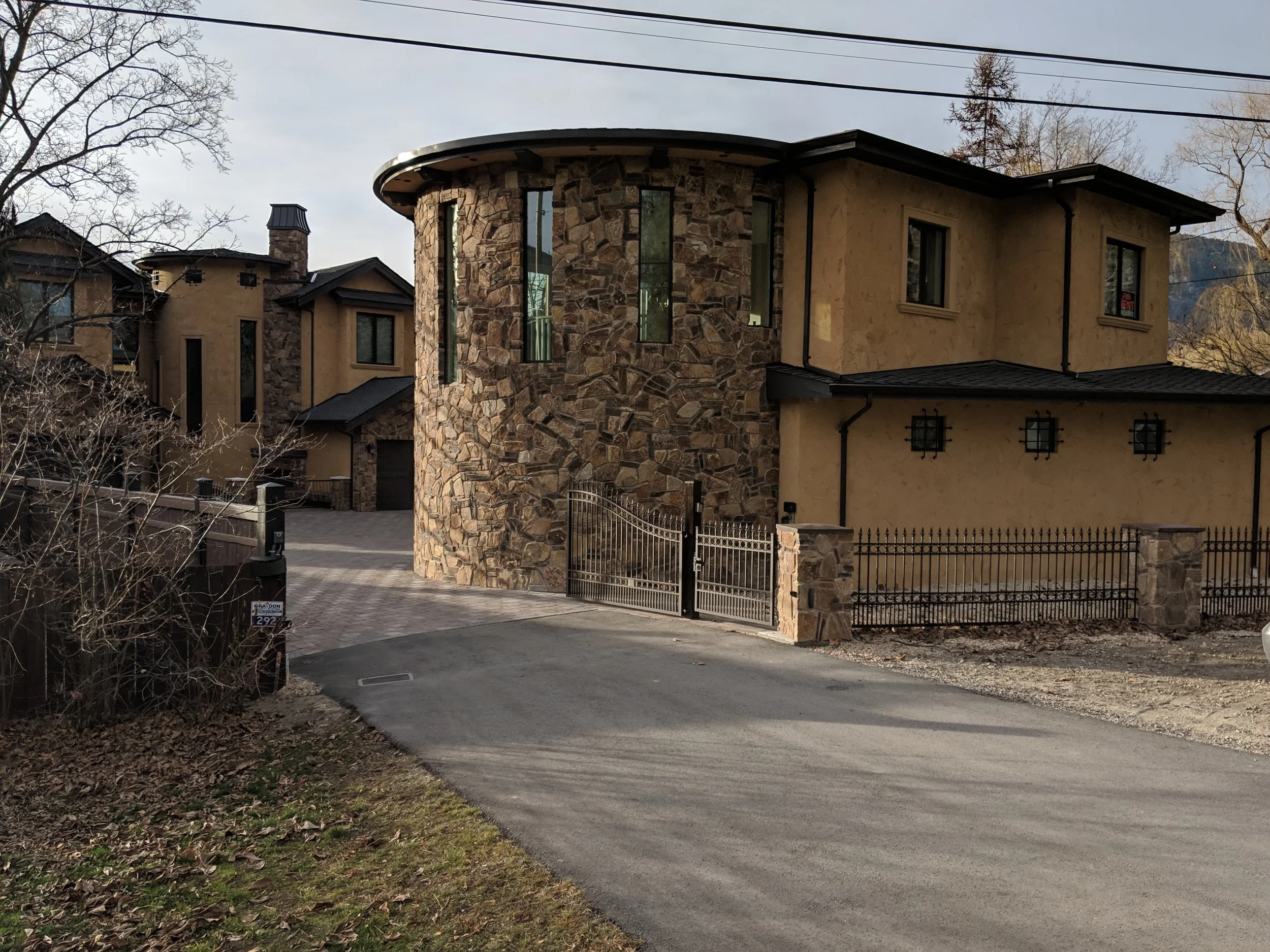 A residential street with a driveway leading to a house that has a rounded stone tower and beige stucco walls. There are leafless trees and power lines visible in the background, suggesting late fall or winter.