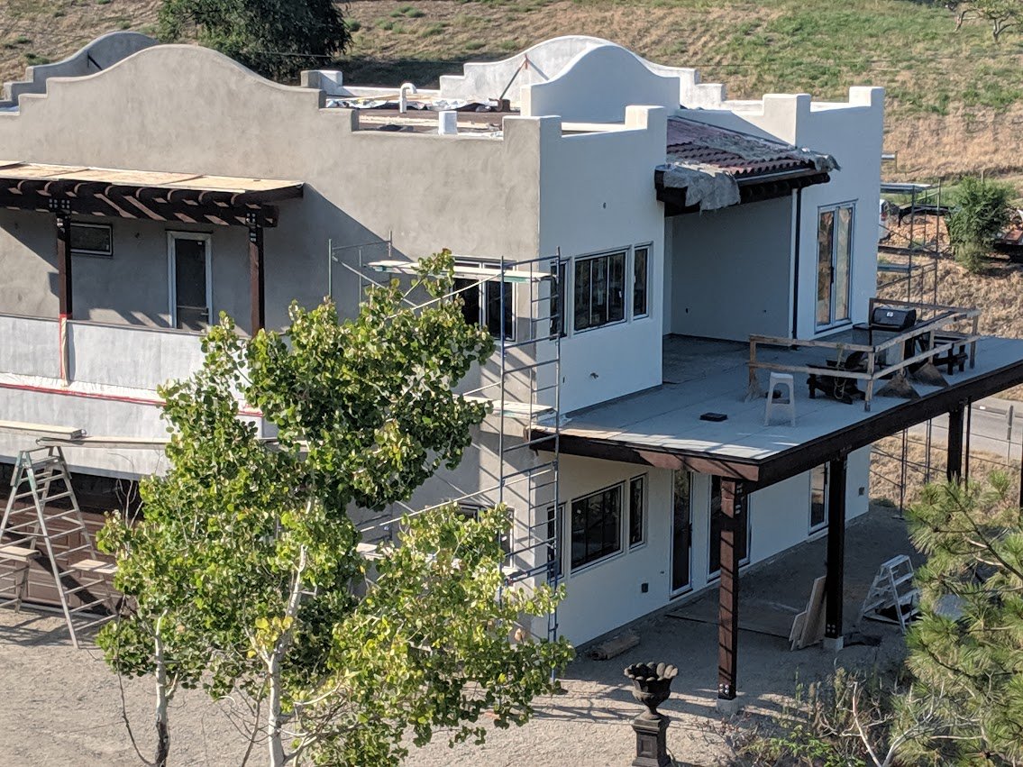 Two-story house under construction with scaffolding, scaffolding, and construction tools visible, surrounded by trees and a dirt ground.