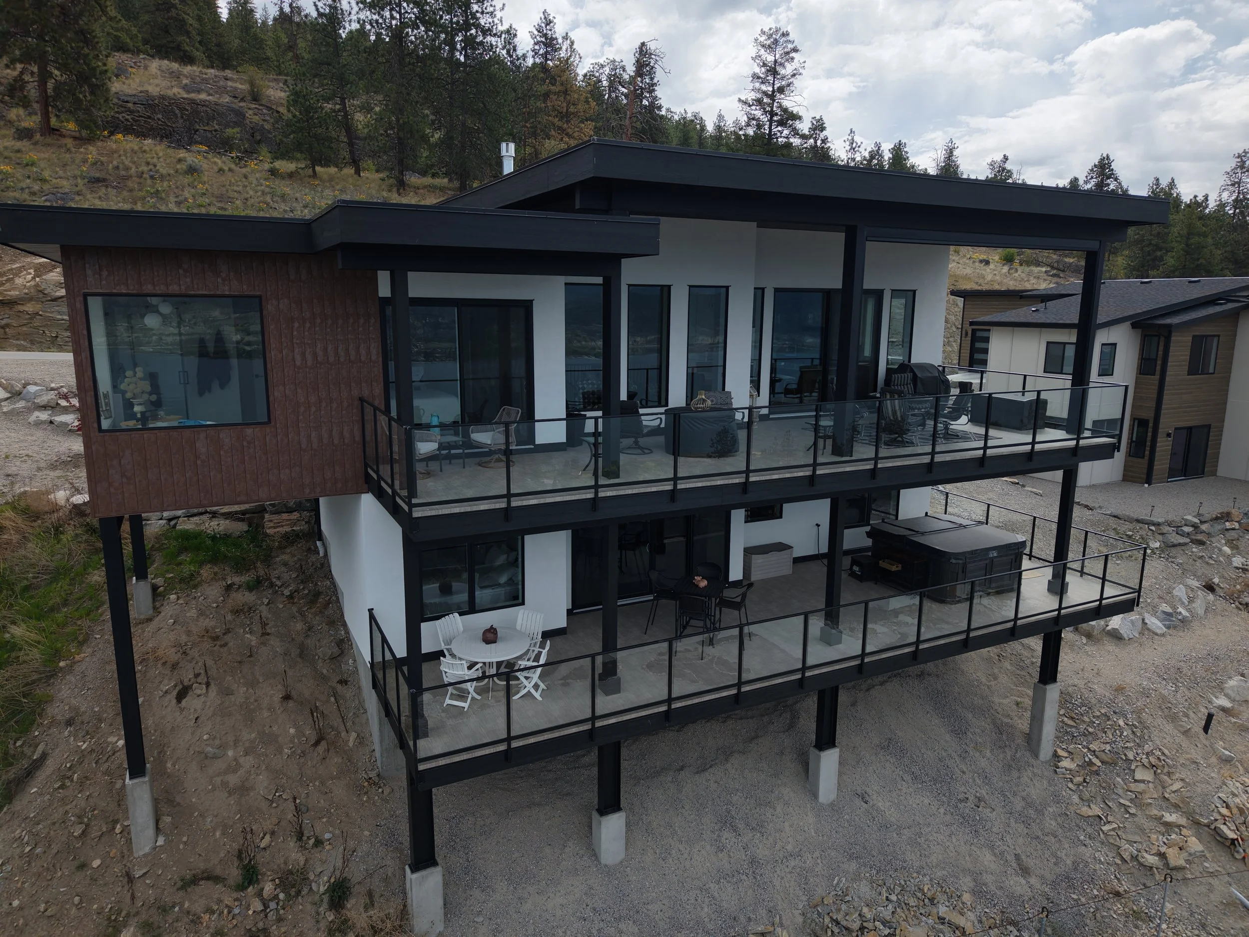 Photo of a modern multi-story house with glass balconies, situated on a hillside with rocky and wooded terrain. The house features black and wood exterior accents, large windows, and outdoor furniture on the balconies. The sky above is partly cloudy.