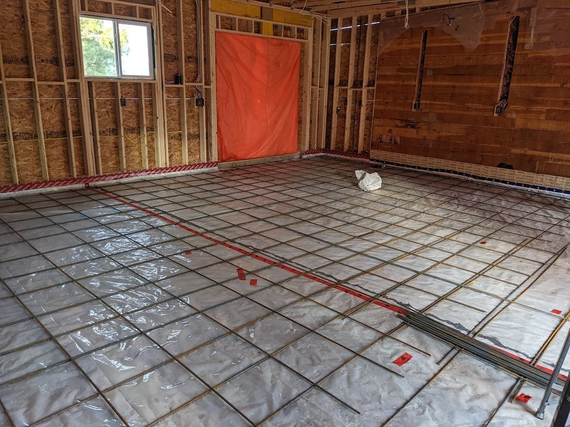 Interior view of a house under construction with exposed wall framing, a window, and a concrete slab floor with a grid of embedded rebar for pouring a concrete floor or foundation.