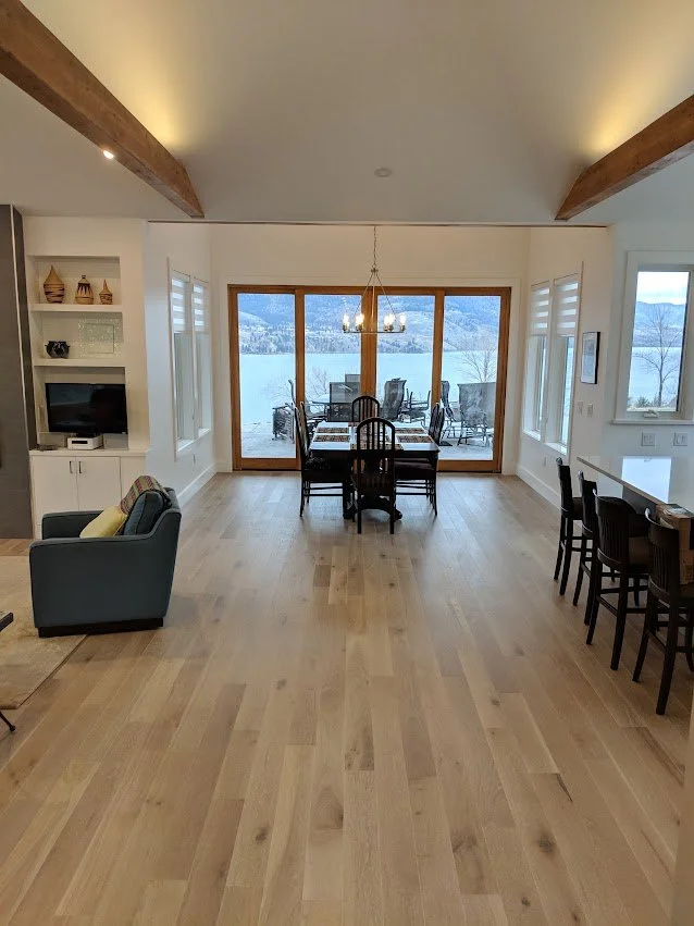 Spacious dining and living area with large glass doors leading to an outdoor patio overlooking a lake and mountains, hardwood floors, and modern furniture.