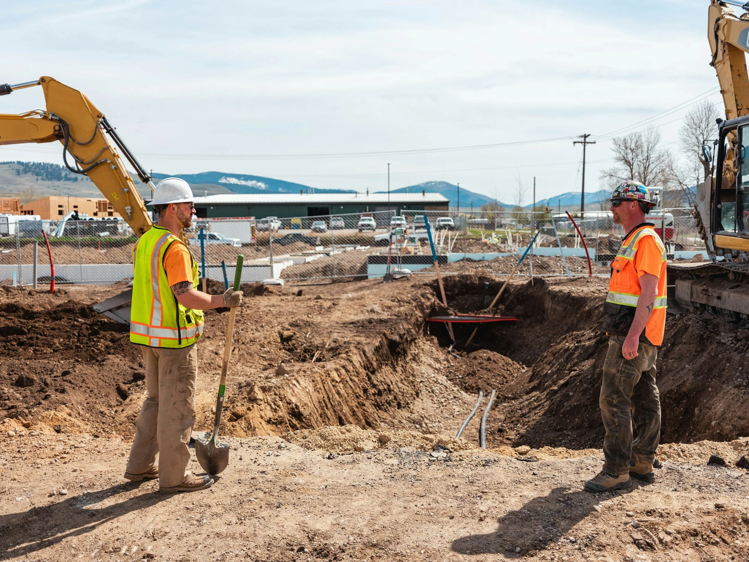 Construction workers in safety vests and helmets standing at a construction site with heavy machinery and excavated earth, mountains in the background.
