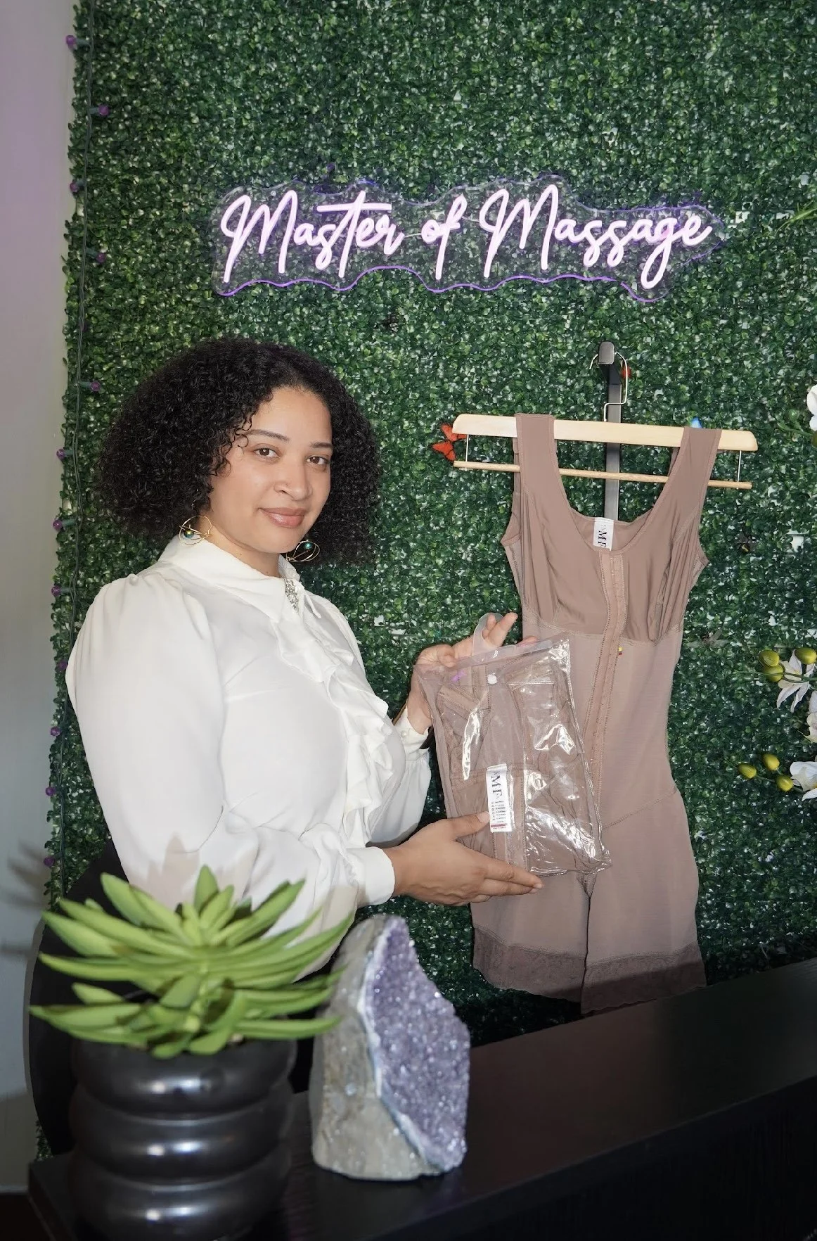 A woman standing at a reception desk holding a clear plastic bag, with a dress on a hanger and a neon sign that reads 'Master of Massage' in the background. The wall behind her has a green leafy pattern, and the desk has a plant and a decorative stone.