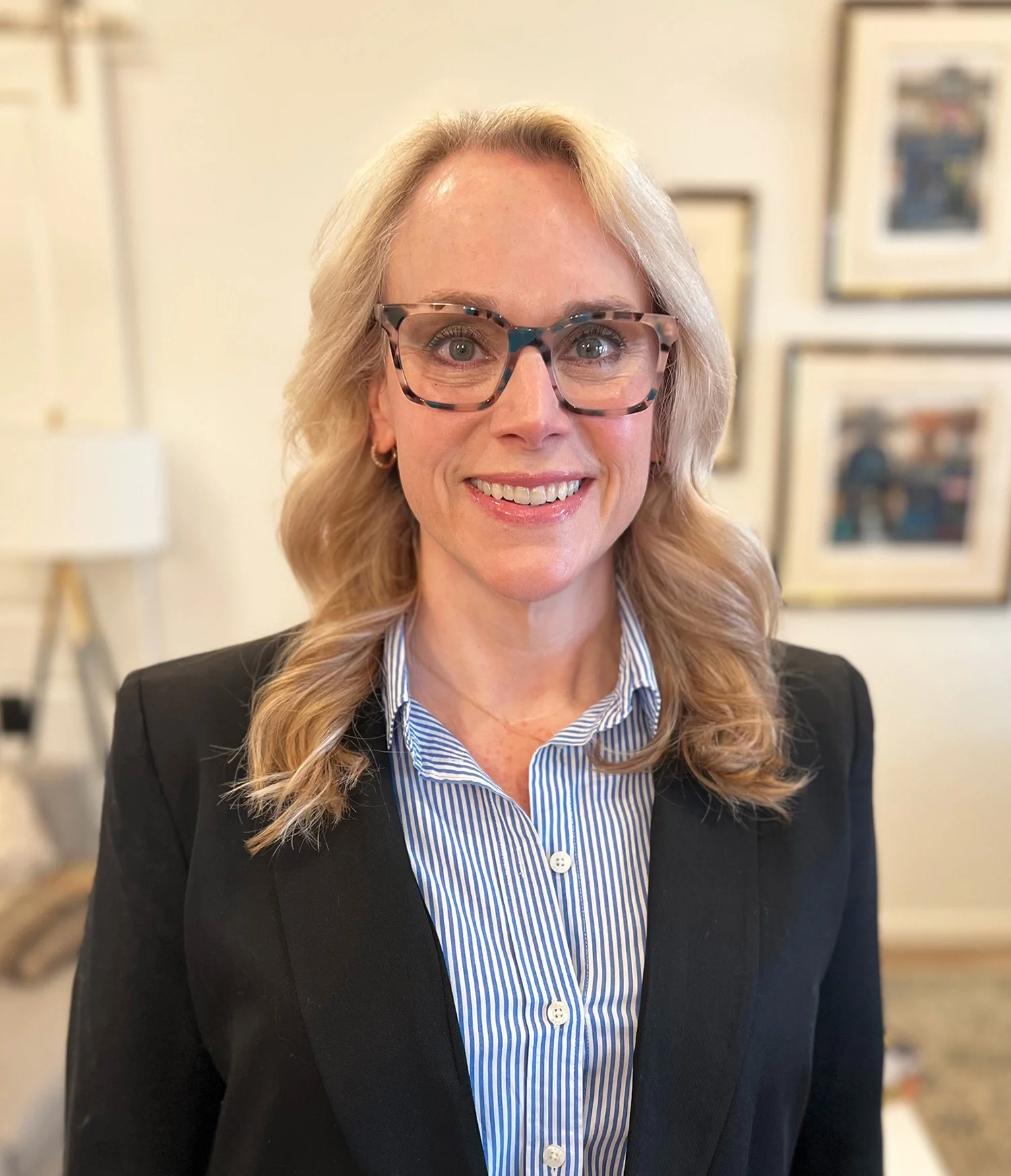 A woman with blonde wavy hair wearing glasses, a striped blue shirt, and a black blazer, smiling in an indoor setting with framed pictures on the wall in the background.