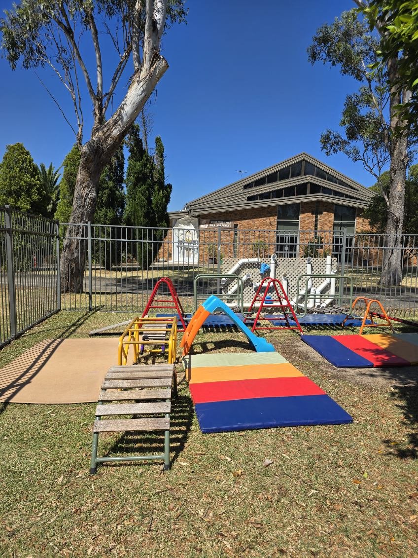 Colorful playground equipment including slides, climbing structures, and mats is enclosed by a metal fence in front of a brick building with a peaked roof, surrounded by trees under a clear blue sky.