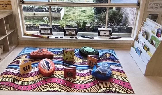 Display of colorful cushions, books, and educational materials on a patterned cloth in front of a window with a view of a garden.