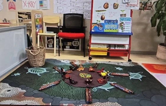 A classroom corner with a circular arrangement of colorful toys on a green and black rug, a black chair with a red cushion, a basket, educational posters, and a red and white two-tiered cart.