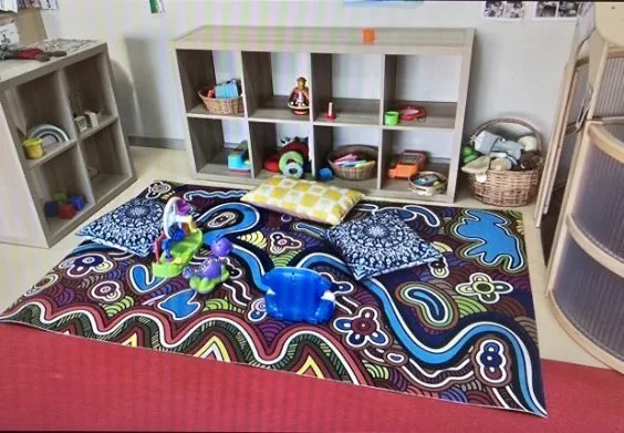 Child's play area with a colorful patterned rug, three pillows, and various toys including stuffed animals and plastic containers, with shelves filled with toys and books in the background.