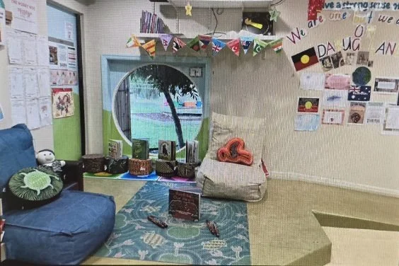 A decorated classroom with a blue couch, a small rug, and a circular window. The wall has colorful flags and artwork, with the words 'We are one' and 'Welcome.' There are books and toys on a yellow shelf and a large cushion on the floor.