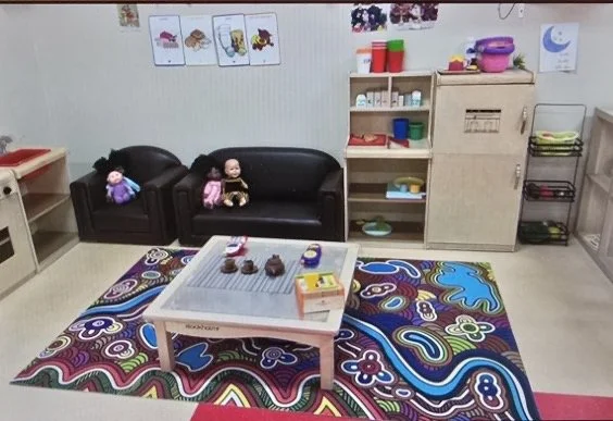 A colorful playroom with a patterned rug, black couch, stuffed dolls, children’s books and toys on shelves, and a wooden cabinet with storage. Posters of cartoon characters on the wall, and a small table with toys and snacks.
