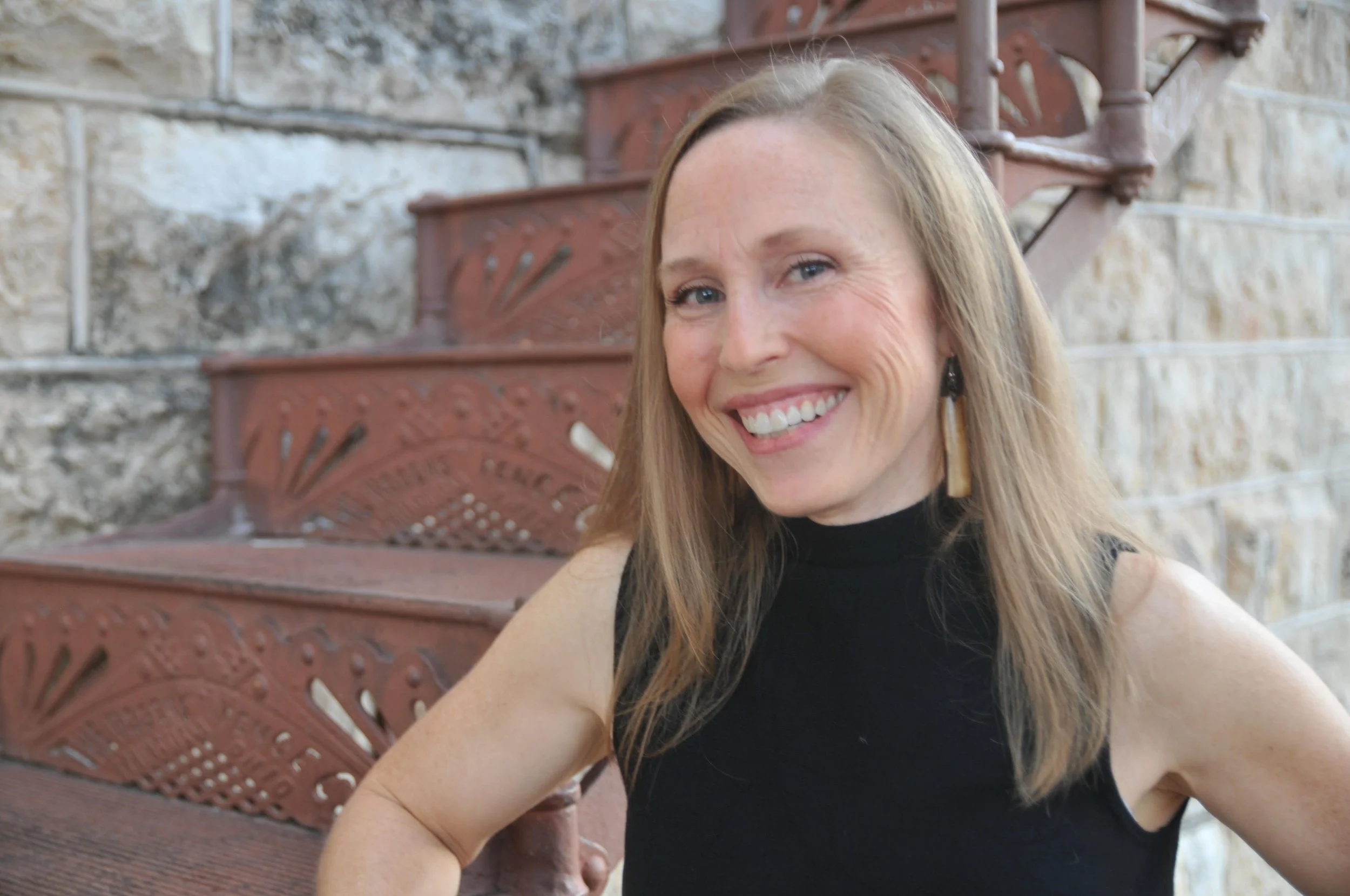 A woman with long blonde hair wearing a sleeveless black top and large earrings, smiling and sitting on red metal stairs next to a stone wall.