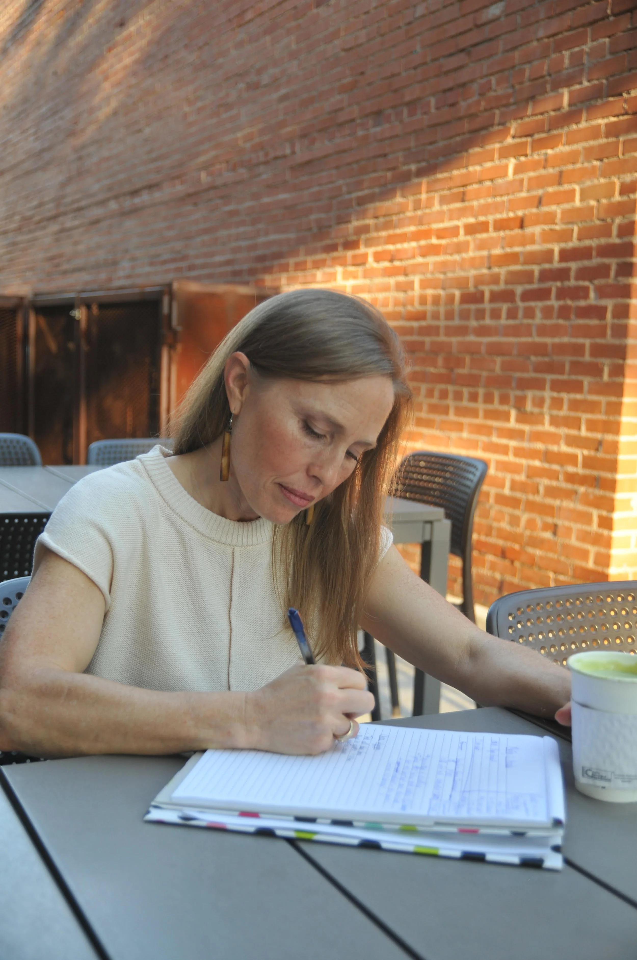 A woman sitting at an outdoor table writing in a notebook with a pen, near a brick wall, with a cup of dessert or yogurt on the table.
