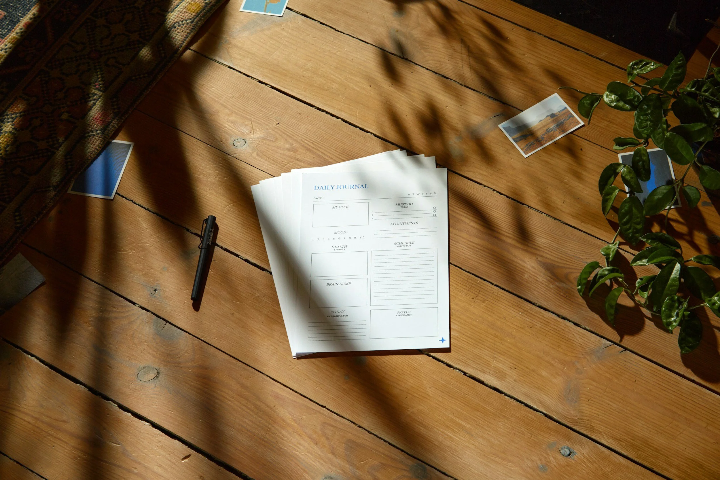 A stack of daily journal planner sheets, a black pen, some loose photographs, and a green leafy plant on a wooden table, with sunlight casting shadows.