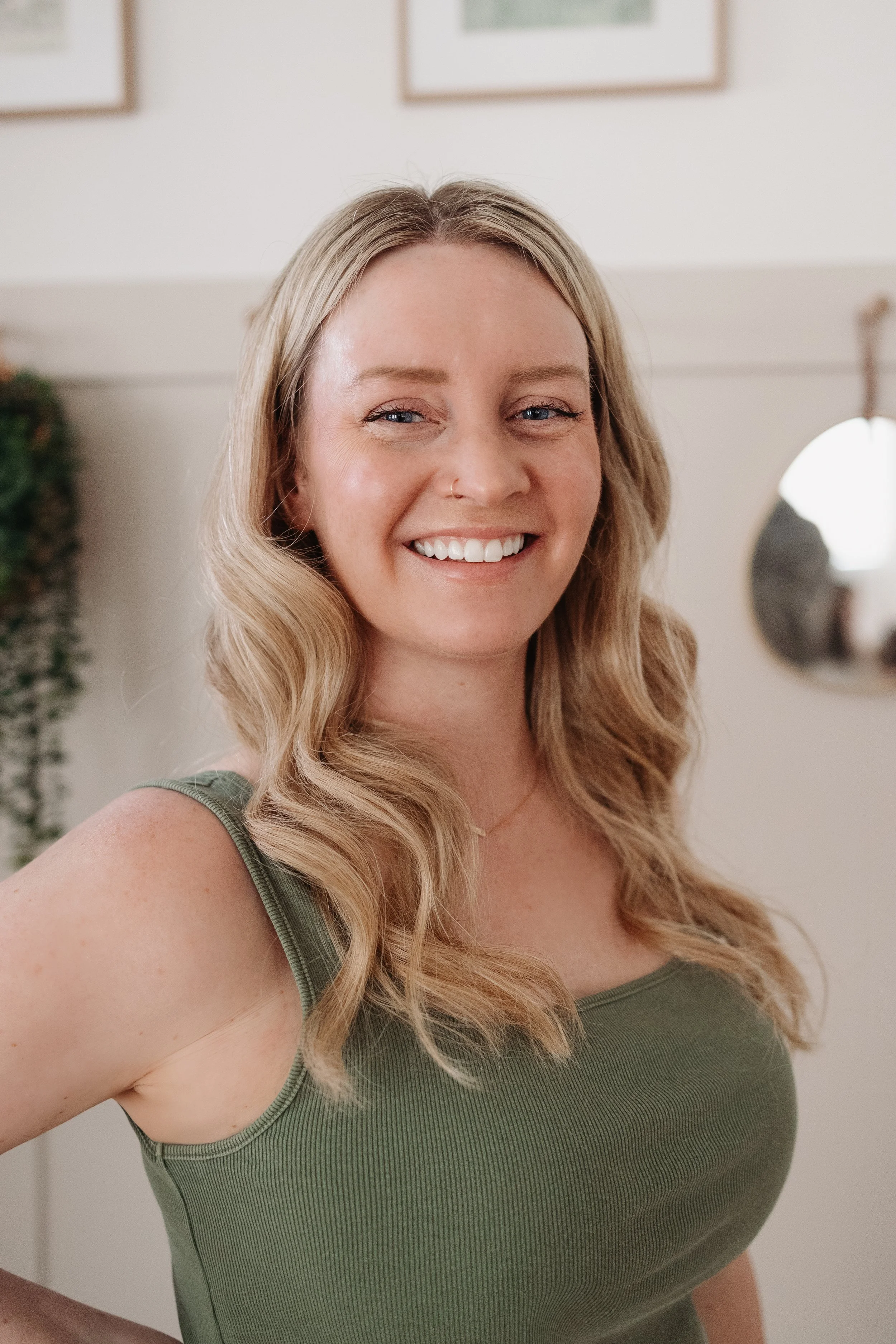 A smiling woman with wavy blonde hair in a green tank top standing indoors with framed pictures on the wall behind her.