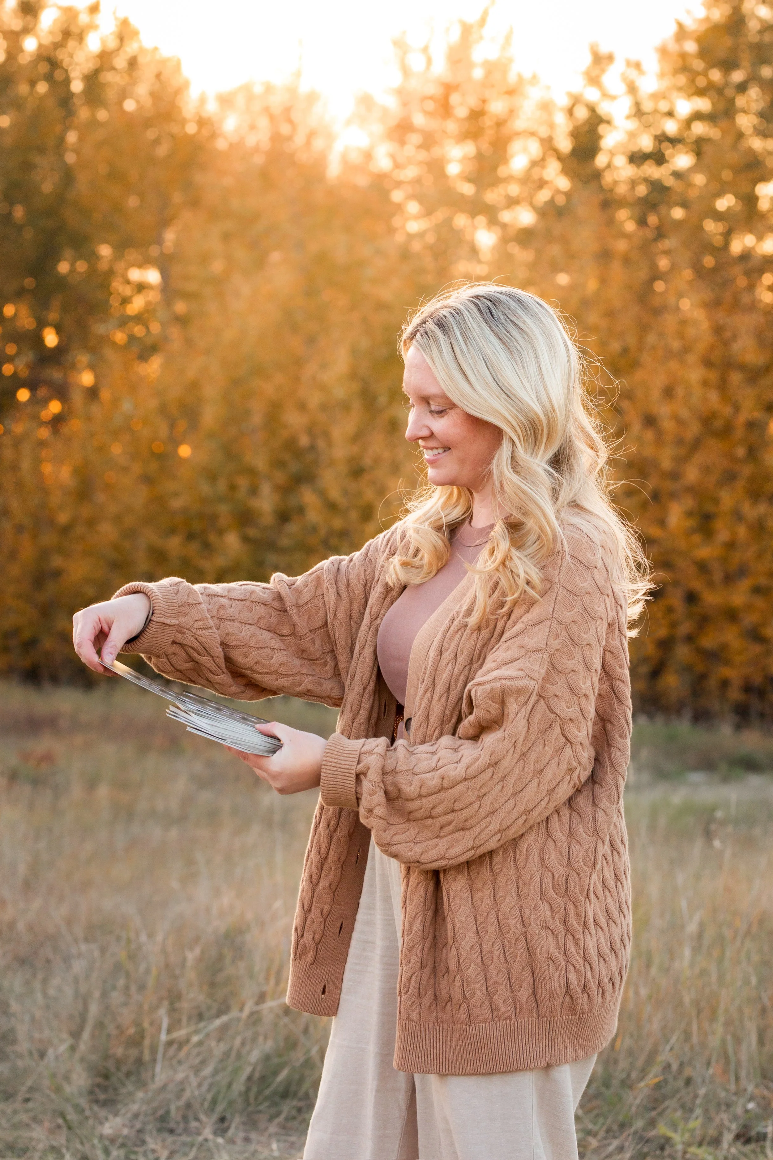A woman with blonde hair wearing a tan sweater and beige pants, smiling while looking at and holding a stack of photo prints in an outdoor setting during sunset, with autumn trees in the background.